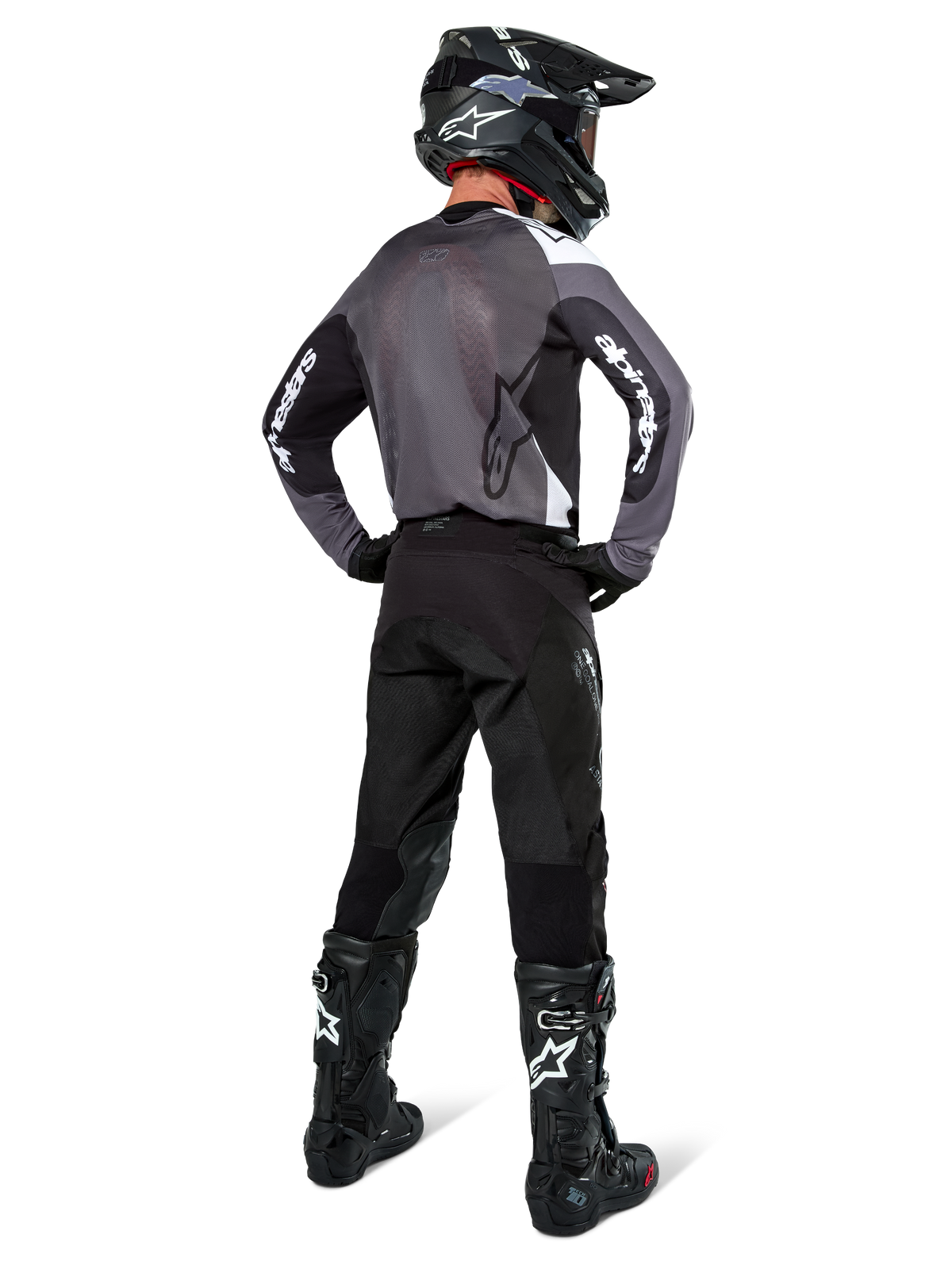 Piloto de motocross con el Jersey Alpinestars Pro-Dura en Black Magnet White, vista trasera que muestra el panel de malla gris y mangas negras con la marca Alpinestars en blanco, coordinado con pantalones negros, casco negro y botas blancas y negras, equipo profesional de enduro