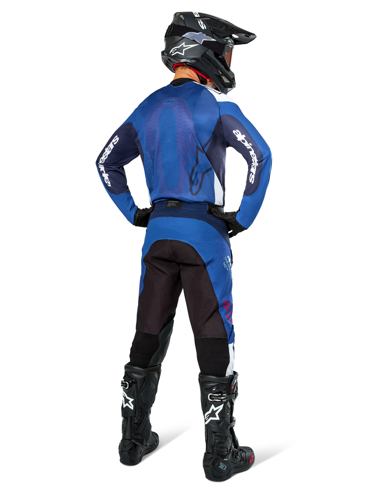 Piloto de motocross visto desde atrás con el Jersey Alpinestars Pro-Dura en azul marino y azul brillante, pantalones azules y negros a juego, casco de motocross negro con gafas y botas negras, conjunto de equipamiento coordinado para carreras todoterreno