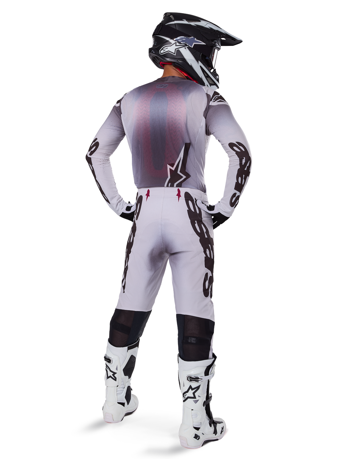 Piloto de motocross visto desde atrás con pantalones Alpinestars 2025 Supertech Maker en plata y negro, jersey de manga larga a juego, casco negro y blanco, y botas blancas y negras, con logotipos negros llamativos en las piernas, equipamiento profesional de carreras off-road