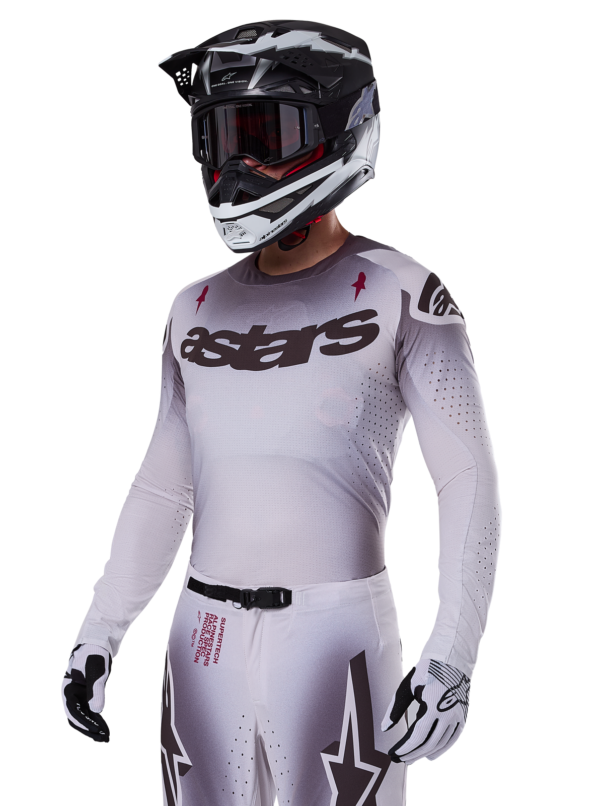 Piloto de motocross con el conjunto de equipamiento Alpinestars 2025 Supertech Maker en blanco, con jersey con degradado gris, logotipo astars en negro y perforaciones cortadas con láser, pantalones a juego con hebilla magnética y gráficos de estrellas, usado con casco y guantes en blanco y negro para un look completo de competición off-road.