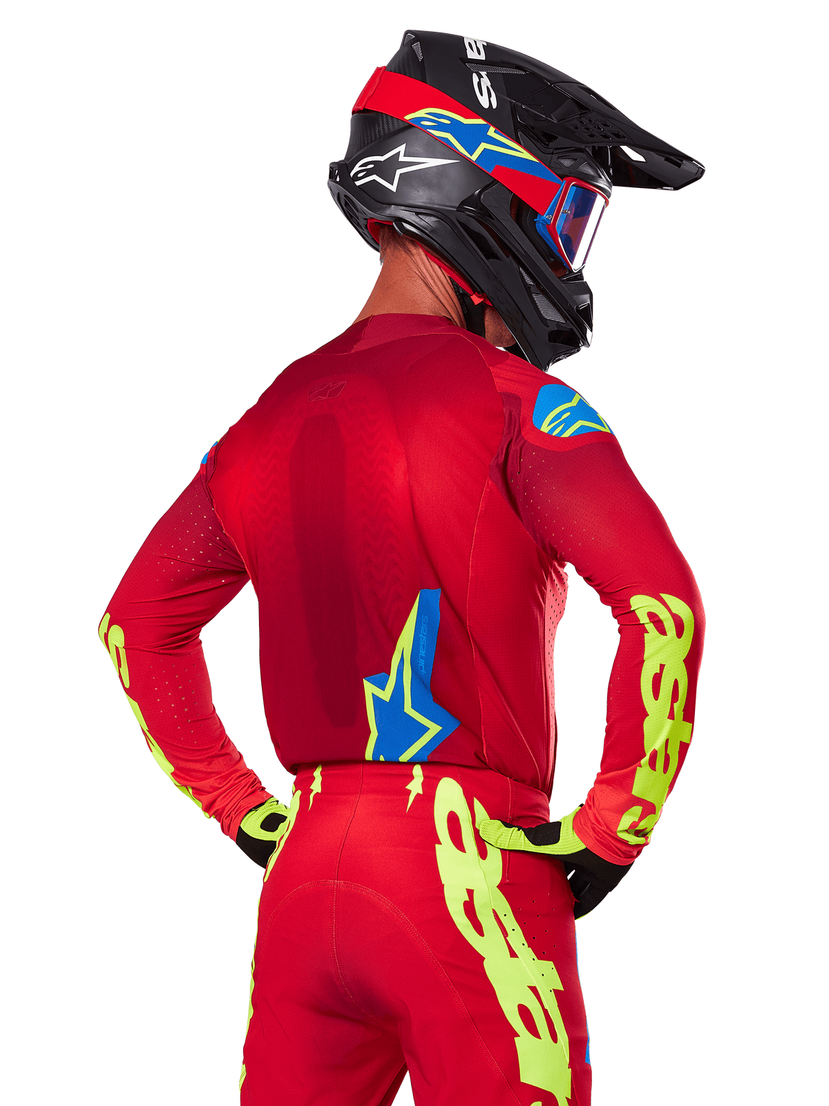 Vista trasera de un piloto de motocross con el conjunto Alpinestars Supertech Maker 2025 en rojo, que incluye un jersey rojo con logotipos en amarillo neón en las mangas, pantalones rojos a juego, un casco negro con detalles en azul y rojo, y guantes coordinados en negro y amarillo.