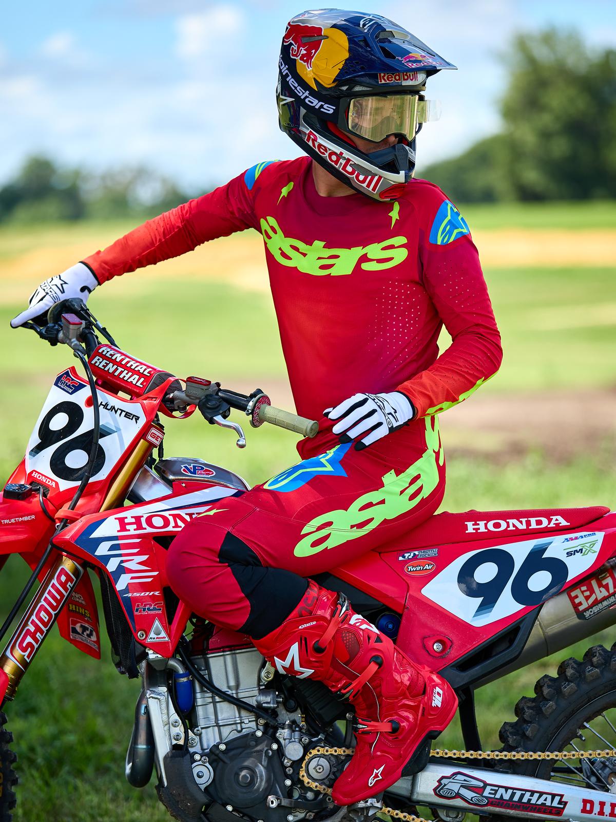 Piloto de motocross en una motocicleta Honda roja vistiendo el conjunto de equipo Alpinestars Supertech Maker 2025 en color rojo, con un jersey rojo con logotipo 'astars' en verde neón y acentos azules en los hombros, pantalones rojos a juego, botas rojas, guantes blancos y casco de Red Bull, kit de carreras profesional para rendimiento todoterreno