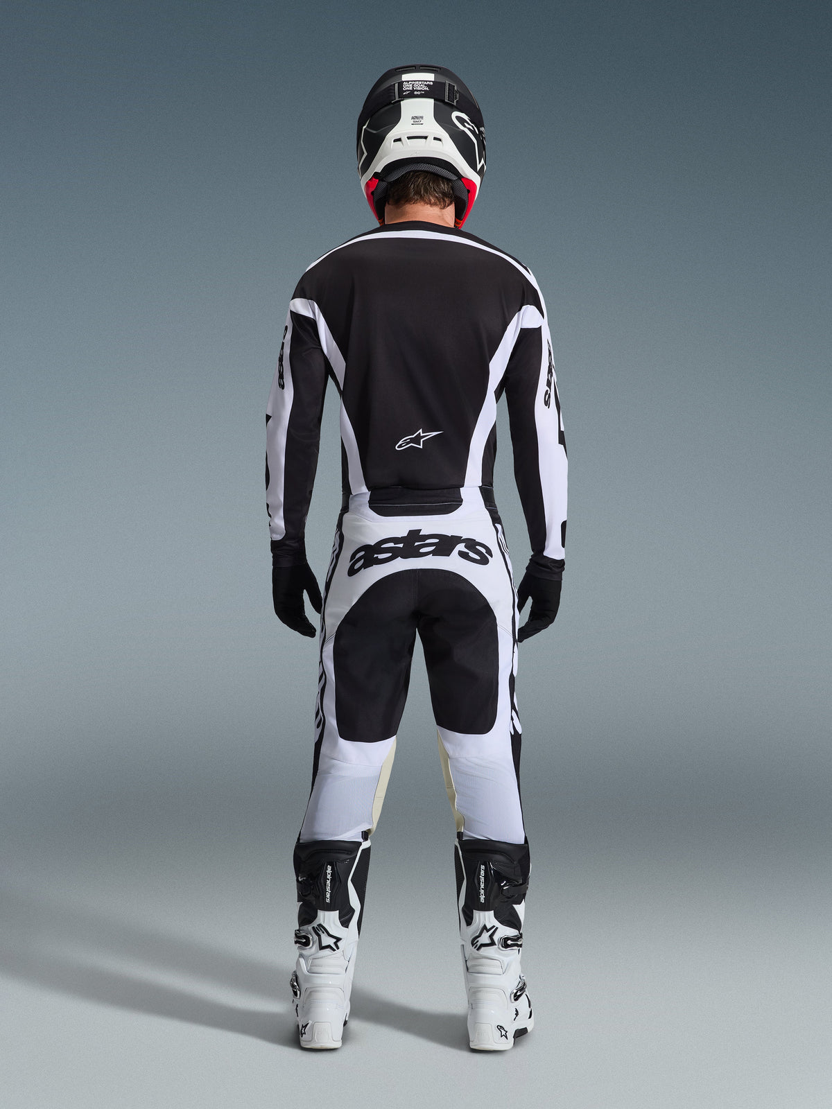 Vista trasera de un piloto de motocross con el conjunto de equipamiento 2026 Racer Riway en blanco y negro, con un jersey negro con detalles blancos, pantalones blancos con el llamativo logotipo 'astars' en la parte trasera, casco negro, botas blancas y guantes negros, equipo profesional completo para carreras off-road.
