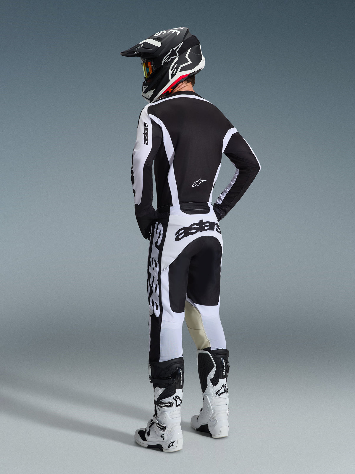 Piloto de motocross desde atrás vistiendo el conjunto Alpinestars 2026 Racer Riway en blanco y negro, con casco negro a juego y botas de motocross blancas, diseño estilizado de alto rendimiento para conducción todoterreno