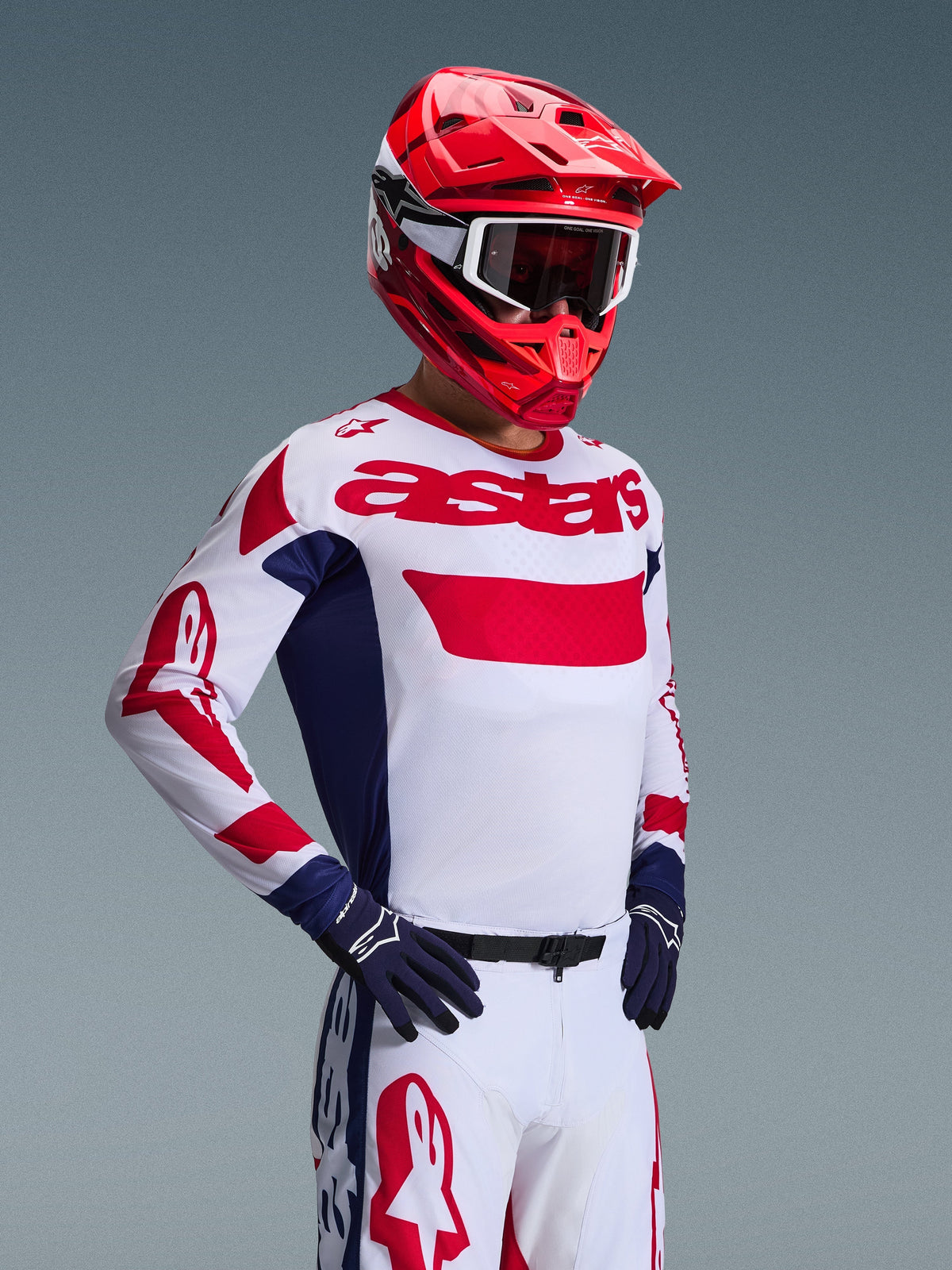 Piloto de motocross con el Jersey Alpinestars 2026 Racer Riway en blanco, azul y rojo, con el logotipo 'astars' en rojo en el pecho y paneles laterales azules, combinado con un casco rojo y pantalones blancos de carreras para un look completo de alto rendimiento off-road.