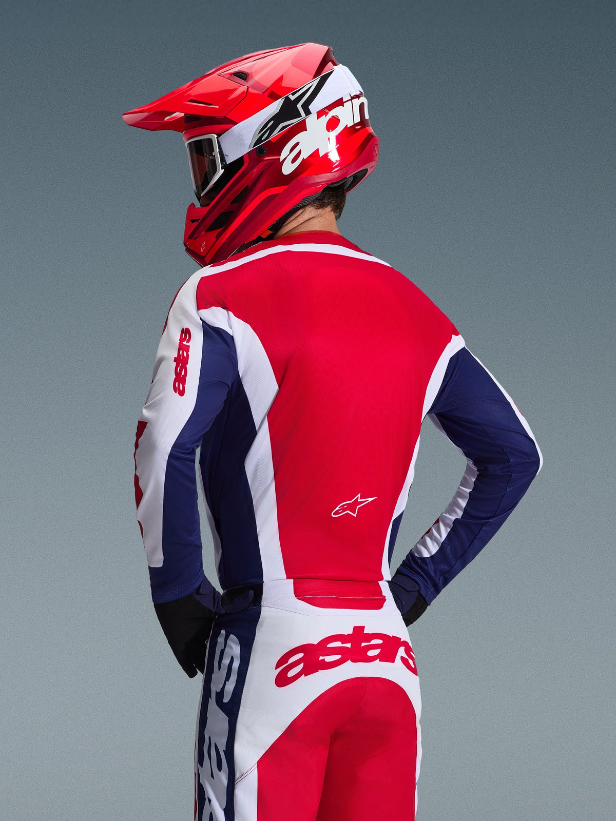Piloto de motocross visto desde atrás con el Jersey Alpinestars Racer Riway 2026, colores blanco, azul y rojo, pantalones de off-road a juego con logotipo astars destacado, casco rojo con logotipo de Alpinestars blanco, guantes negros, equipamiento de motocross coordinado