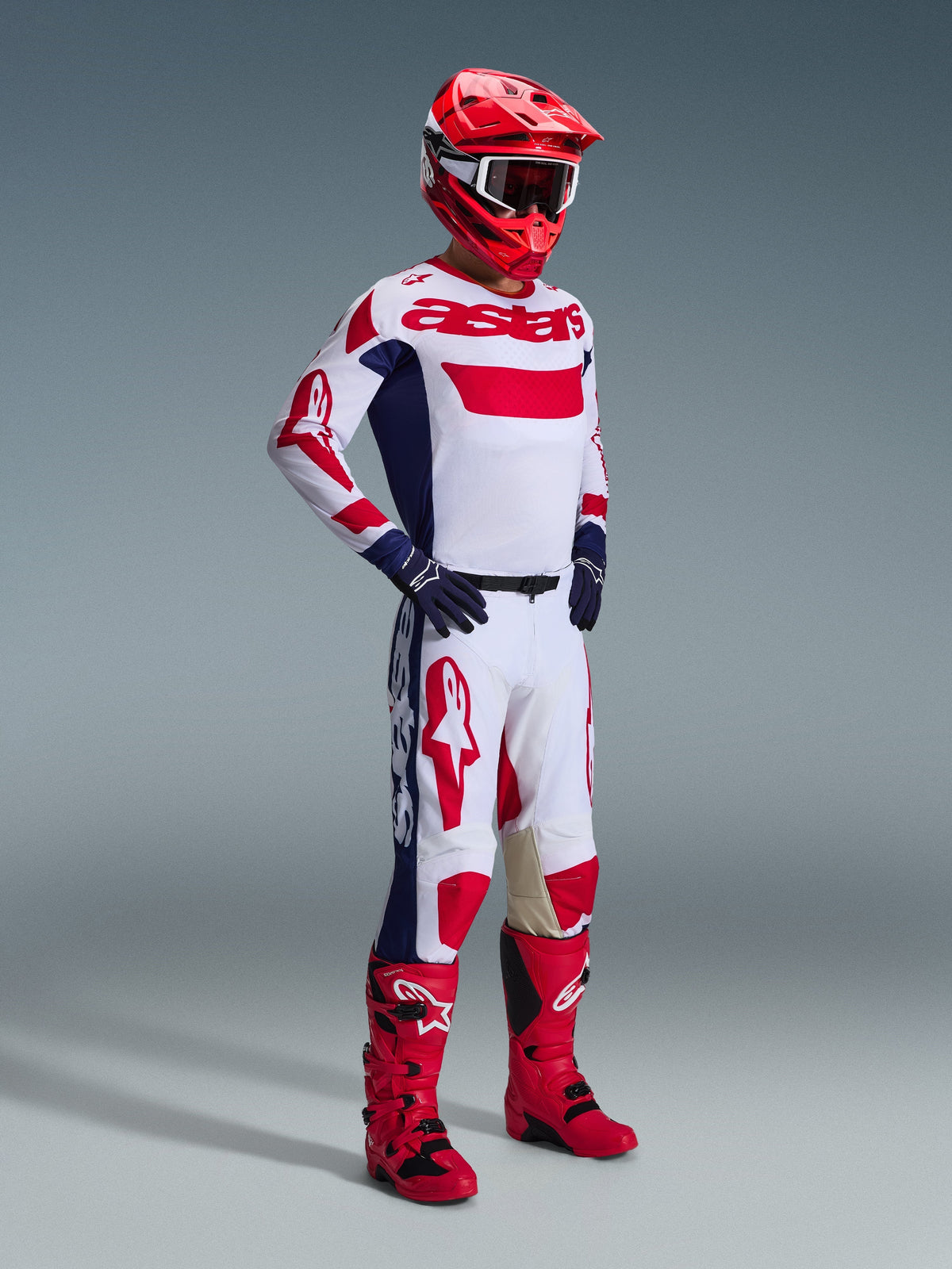 Piloto de motocross con el Jersey Racer Riway 2026 en blanco, azul y rojo, pantalones a juego en blanco y rojo, casco rojo con gafas blancas, guantes azul marino y botas de motocross rojas, conjunto completo de equipamiento de competición para rendimiento off-road.
