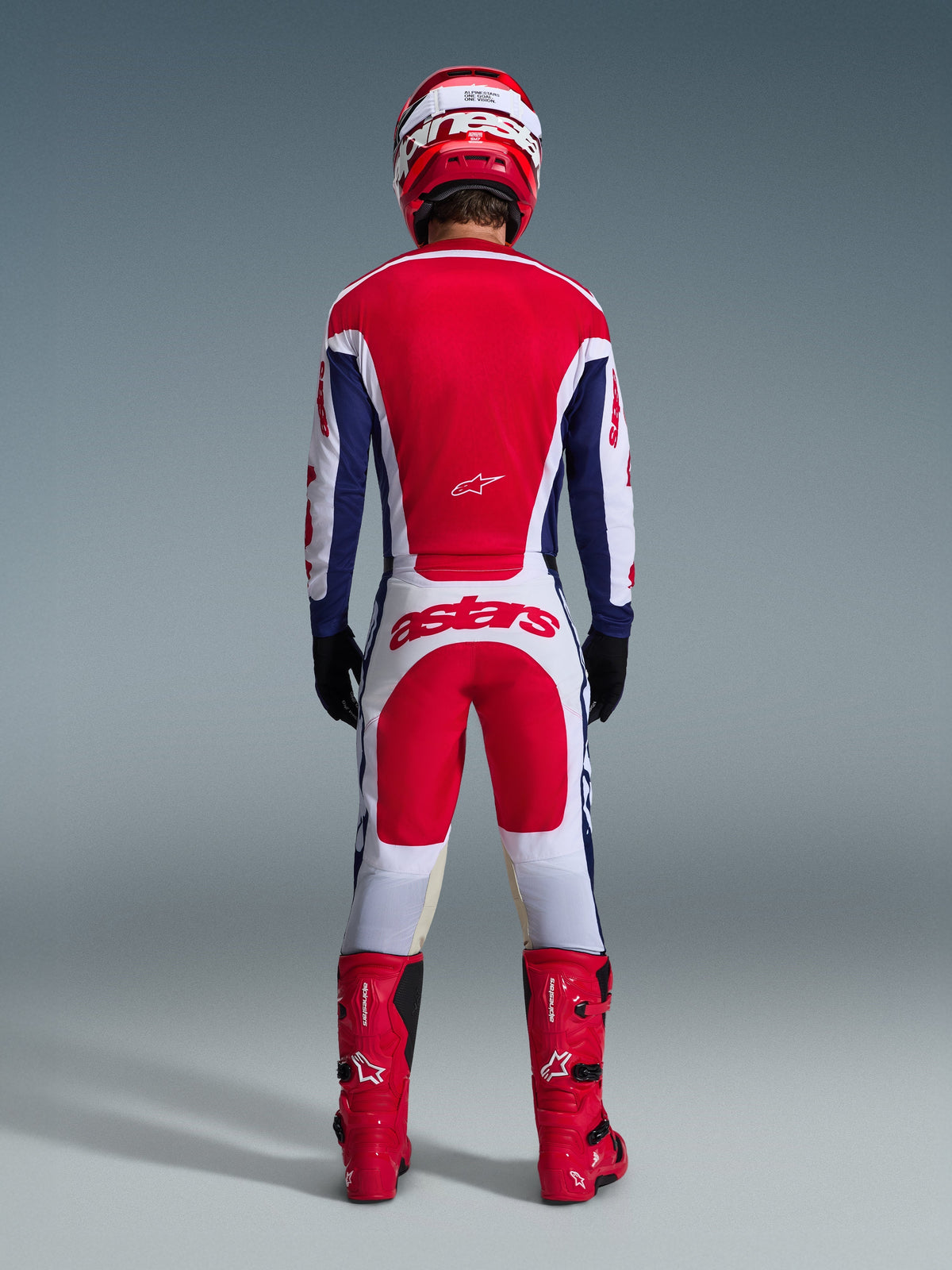 Piloto de motocross con equipamiento de competición completo visto desde atrás, Jersey Alpinestars Racer Riway 2026 en blanco, azul y rojo, pantalones blancos y rojos a juego con el llamativo logotipo 'astars', botas rojas y casco rojo y blanco, conjunto de carreras off-road coordinado.