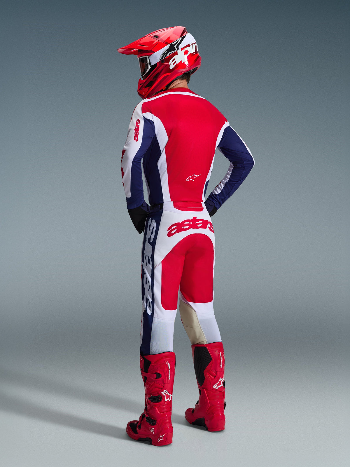 Piloto de motocross visto desde atrás vistiendo el Jersey Alpinestars Racer Riway 2026 en blanco, azul y rojo, pantalones de motocross a juego con la marca 'astars' en el asiento, casco rojo y blanco, y botas de motocross rojas, equipamiento completo de carreras off-road