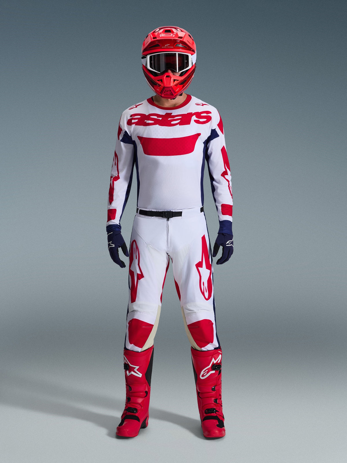 Piloto de motocross con equipamiento completo de competición, Alpinestars 2026 Racer Riway Jersey en blanco, azul y rojo, pantalones blancos y rojos a juego, casco rojo, botas rojas y guantes azul marino, conjunto coordinado para carreras off-road