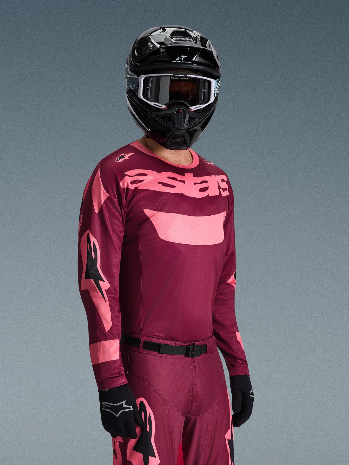 Piloto de motocross con el conjunto de equipamiento Alpinestars 2026 Racer Riway, jersey burdeos y rosa con logotipo 'astars' llamativo, pantalones a juego con detalles rosas, casco y guantes negros, traje de carreras profesional para motocross off-road