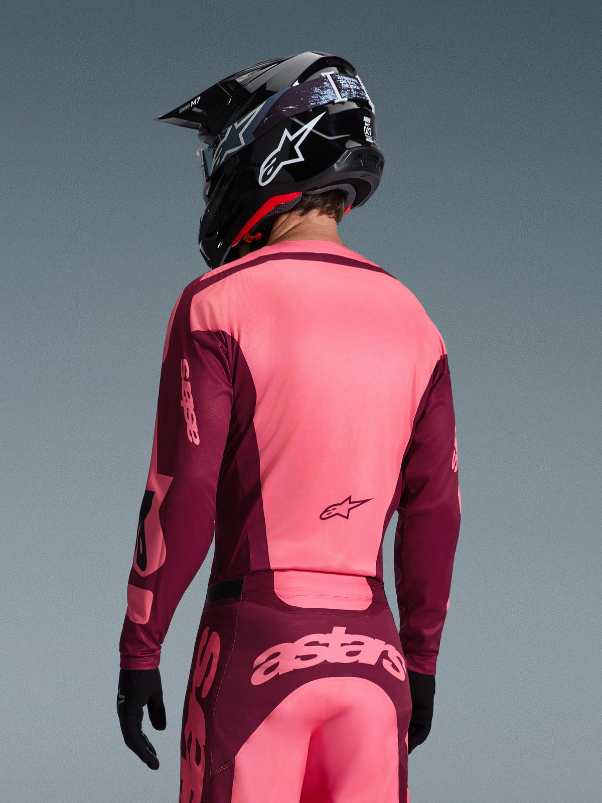 Piloto de motocross visto desde atrás con el conjunto de equipación Alpinestars 2026 Racer Riway, jersey y pantalones en rosa brillante y granate, casco de motocross negro con logo blanco y guantes negros, con el logotipo 'astars' en la parte trasera.