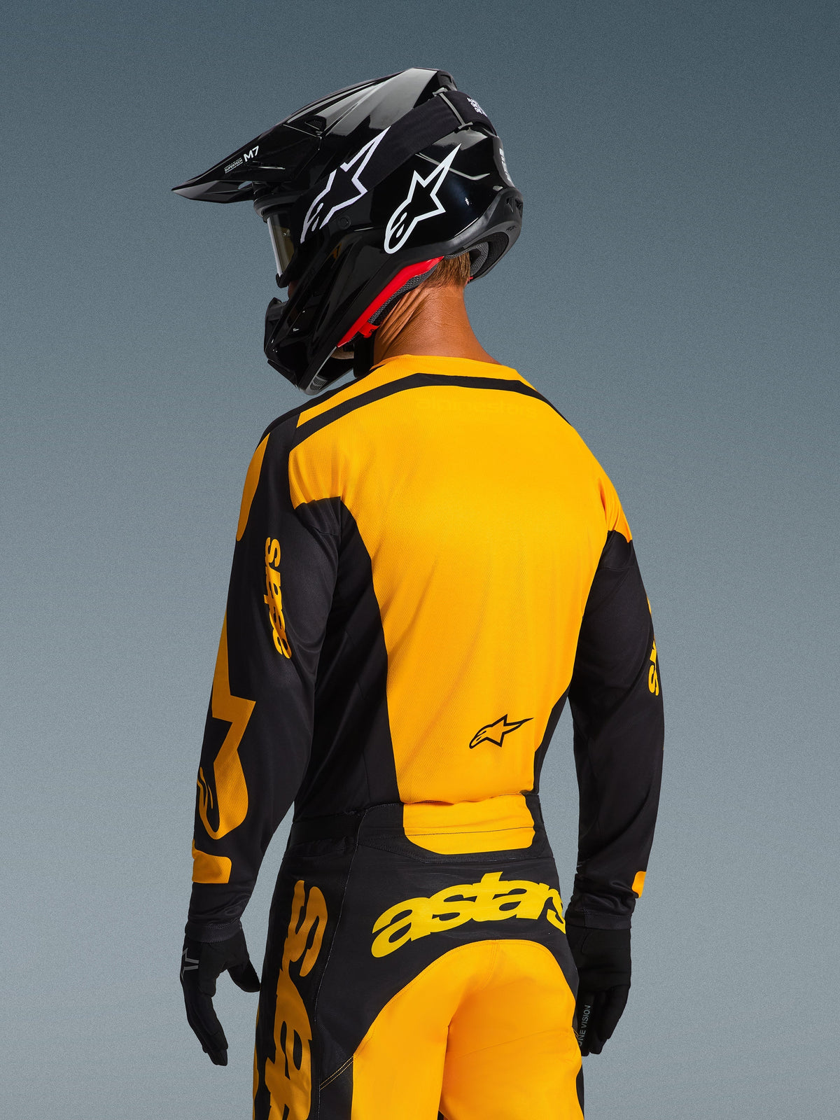 Piloto de motocross visto desde atrás vistiendo el Jersey Racer Riway 2026 y pantalones a juego en amarillo y negro, con logotipos destacados de Alpinestars, casco negro con logos de estrellas blancas y guantes negros; conjunto de equipación de motocross coordinado para carreras off-road.