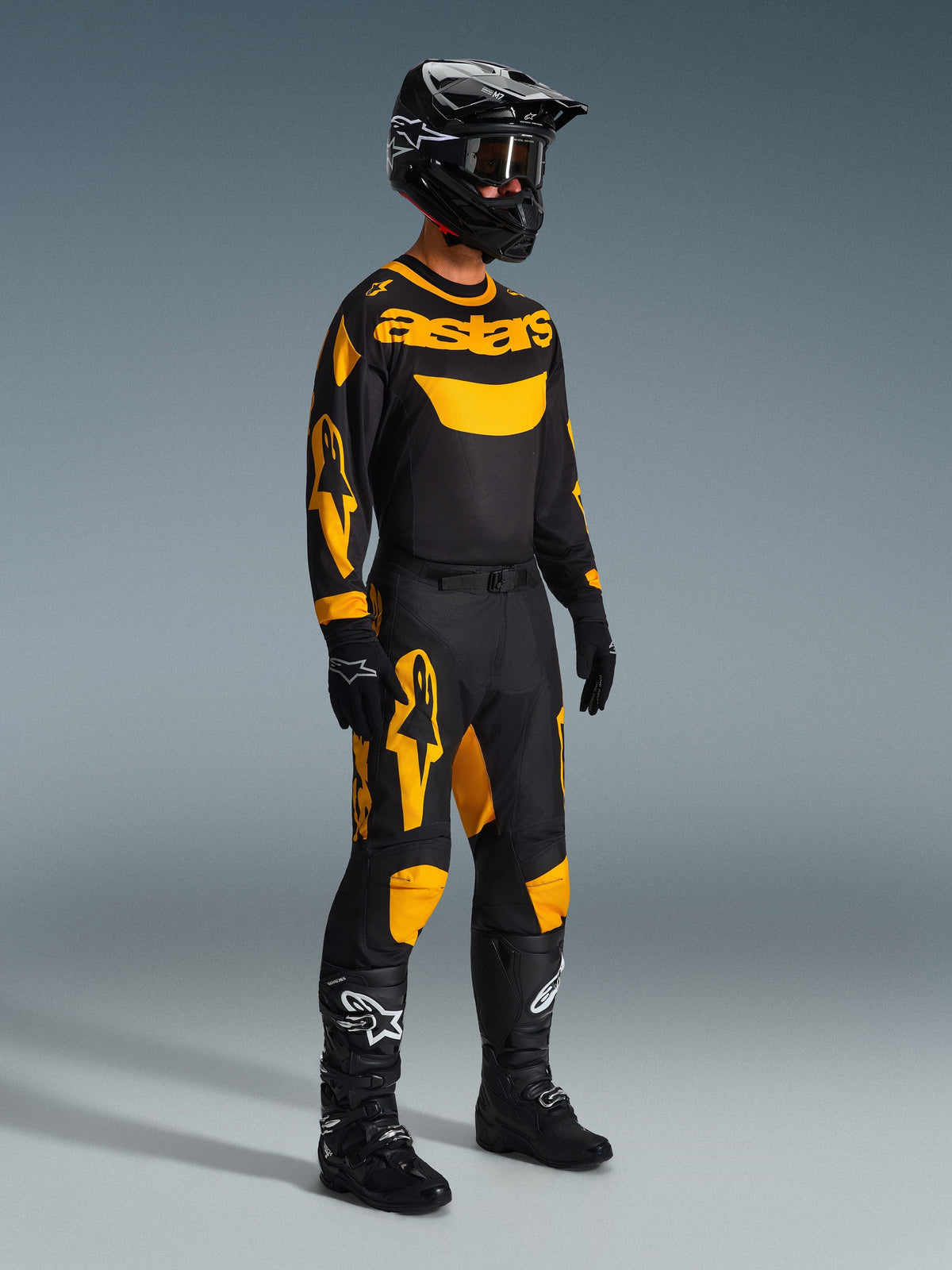 Piloto de motocross con equipamiento completo de competición, jersey Alpinestars 2026 Racer Riway en amarillo y negro, pantalones a juego en negro y amarillo, casco negro con gafas, botas y guantes negros, conjunto coordinado para carreras off-road