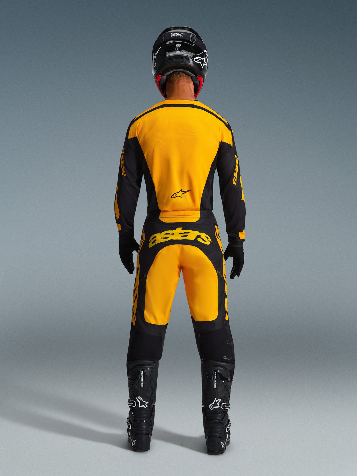 Piloto de motocross visto desde atrás vistiendo el conjunto Alpinestars 2026 Racer Riway, jersey y pantalones amarillos y negros con marca 'astars' en la parte trasera, combinado con casco, guantes y botas negros, equipamiento completo de carreras off-road.