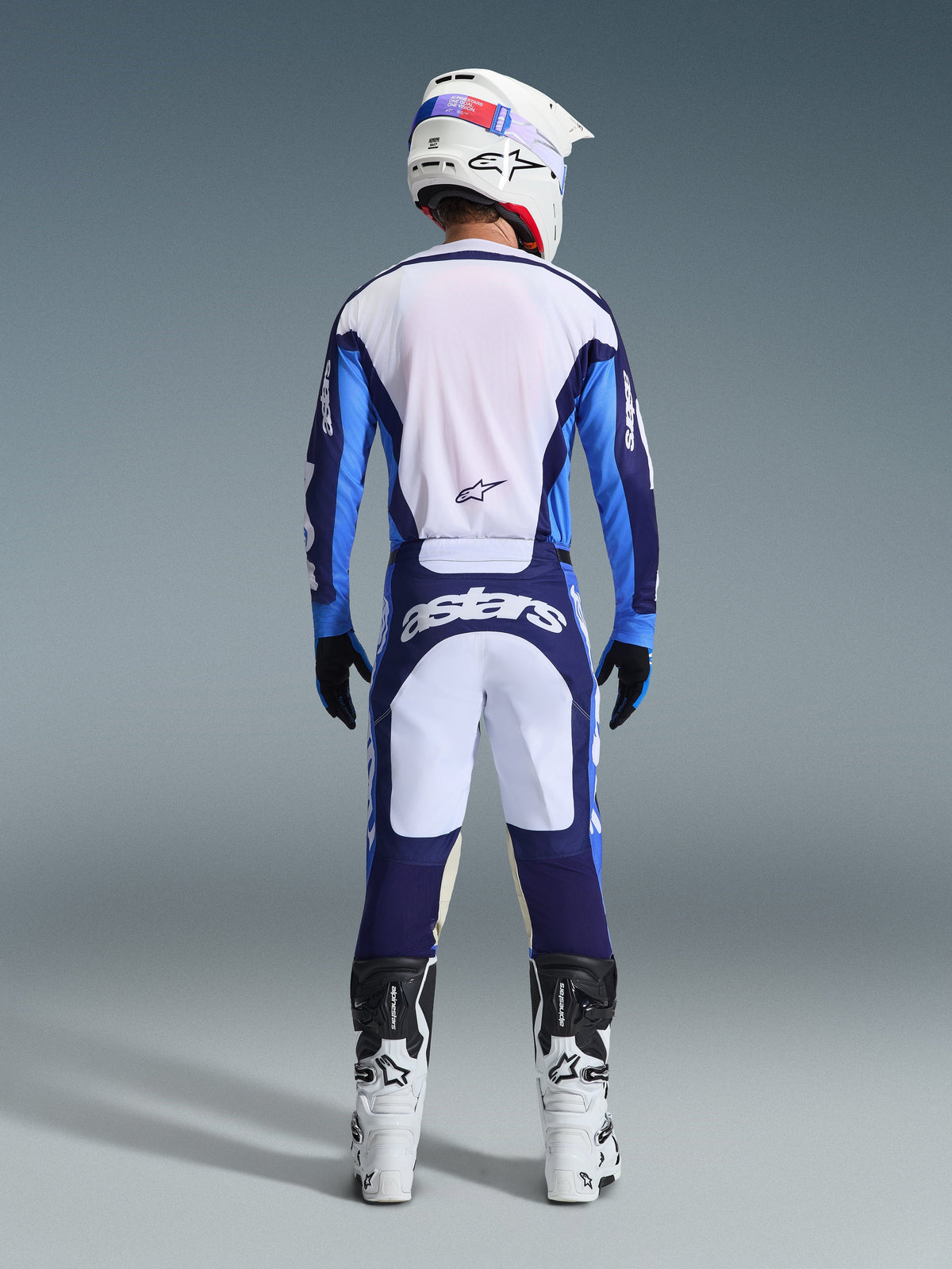 Rear view of a motocross rider wearing the Alpinestars 2026 Racer Riway Jersey and Pants in Ucla Blue and White, coordinated with a white helmet, black gloves, and white motocross boots, high-performance gear for off-road racing