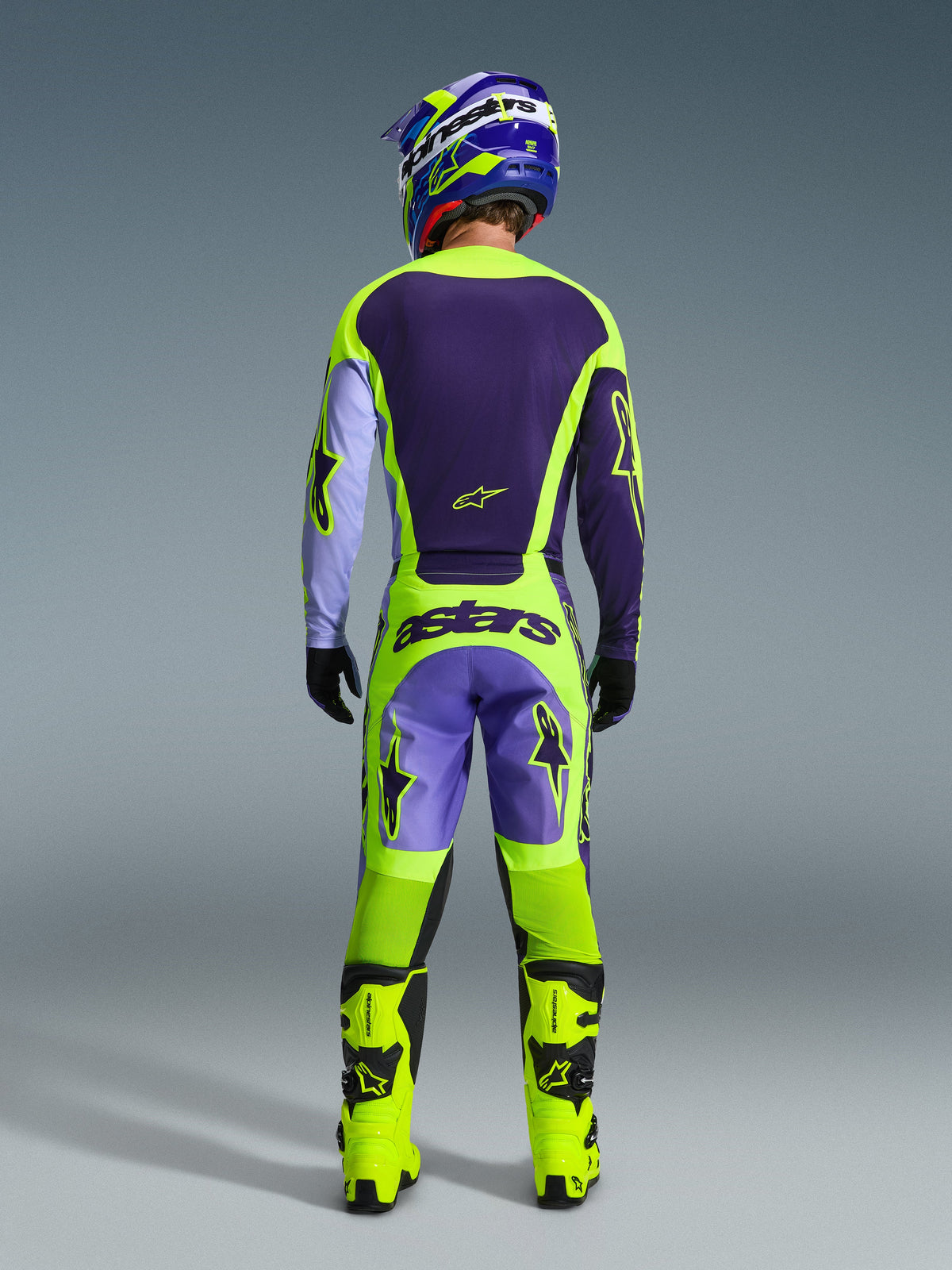 Motocross rider wearing Alpinestars 2026 Racer Portl Gear Set, rear view, Dark Purple and Neon Yellow jersey with Lavender sleeves, matching Purple and Neon Yellow pants with 'astars' branding, bright Neon Yellow boots, professional off-road racing outfit