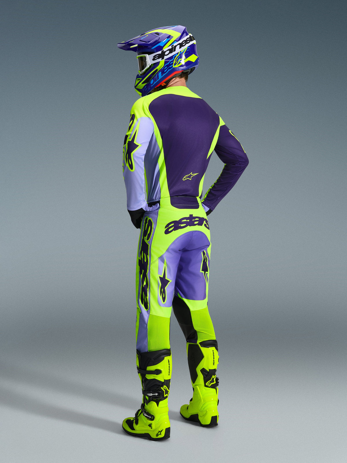 Motocross rider wearing Alpinestars 2026 Racer Portl Gear Set, Purple and Neon Yellow jersey and pants with lavender panels, matching purple and yellow helmet, bright neon yellow boots, rear view showing professional off-road racing gear.