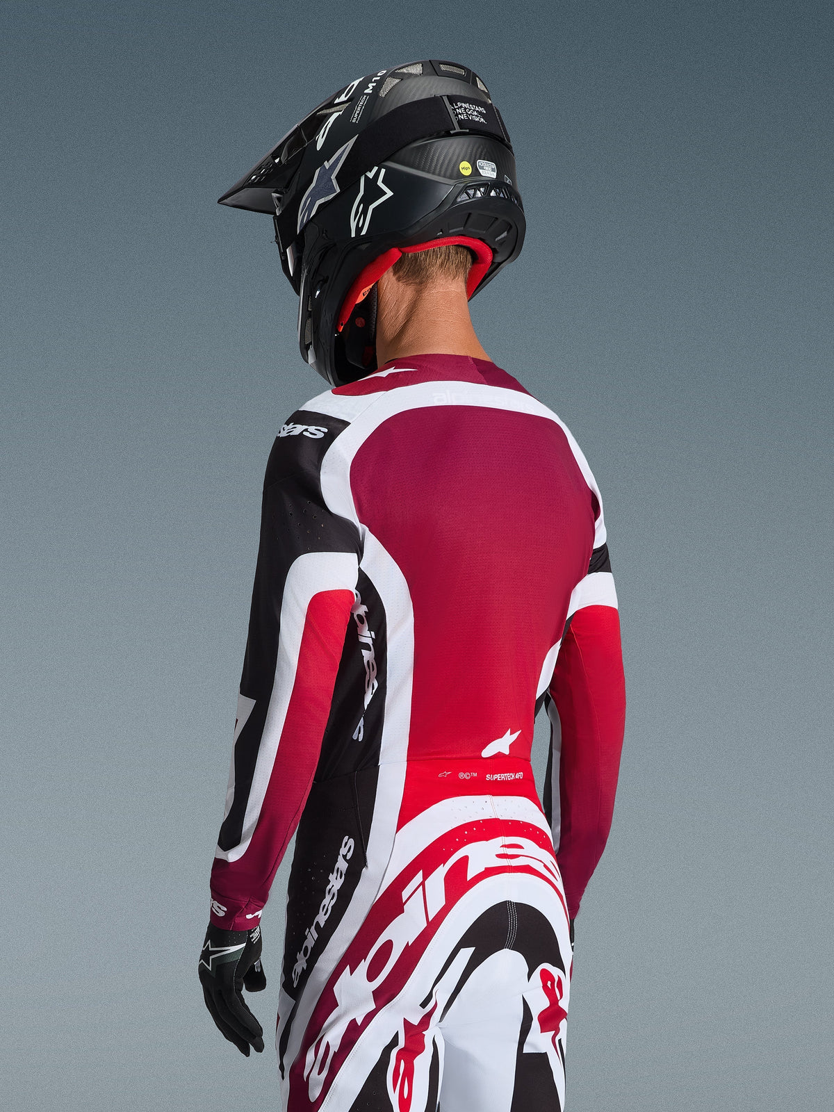 Vista trasera de un piloto de motocross con el Jersey Supertech AFD Vista 2026 en negro, rojo y blanco, combinado con pantalones a juego y un casco negro, con logotipos de Alpinestars destacados, paneles de malla rojos y un ajuste atlético de competición.