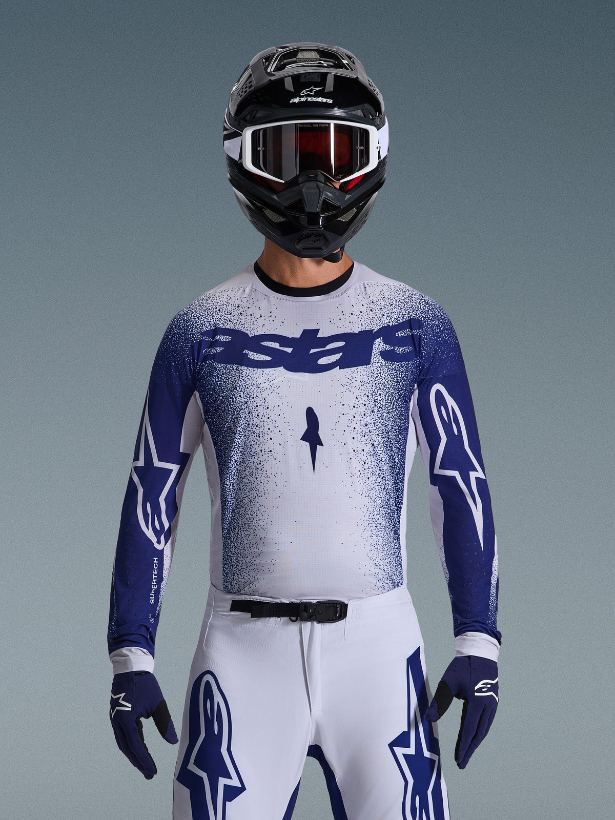 Motocross rider wearing the Alpinestars 2026 Supertech Scenz Gear Set, white jersey with a blue speckled gradient and 'astars' branding, matching white pants with blue accents, black helmet with white goggles, and blue gloves, showing the athletic pro-level racing fit.