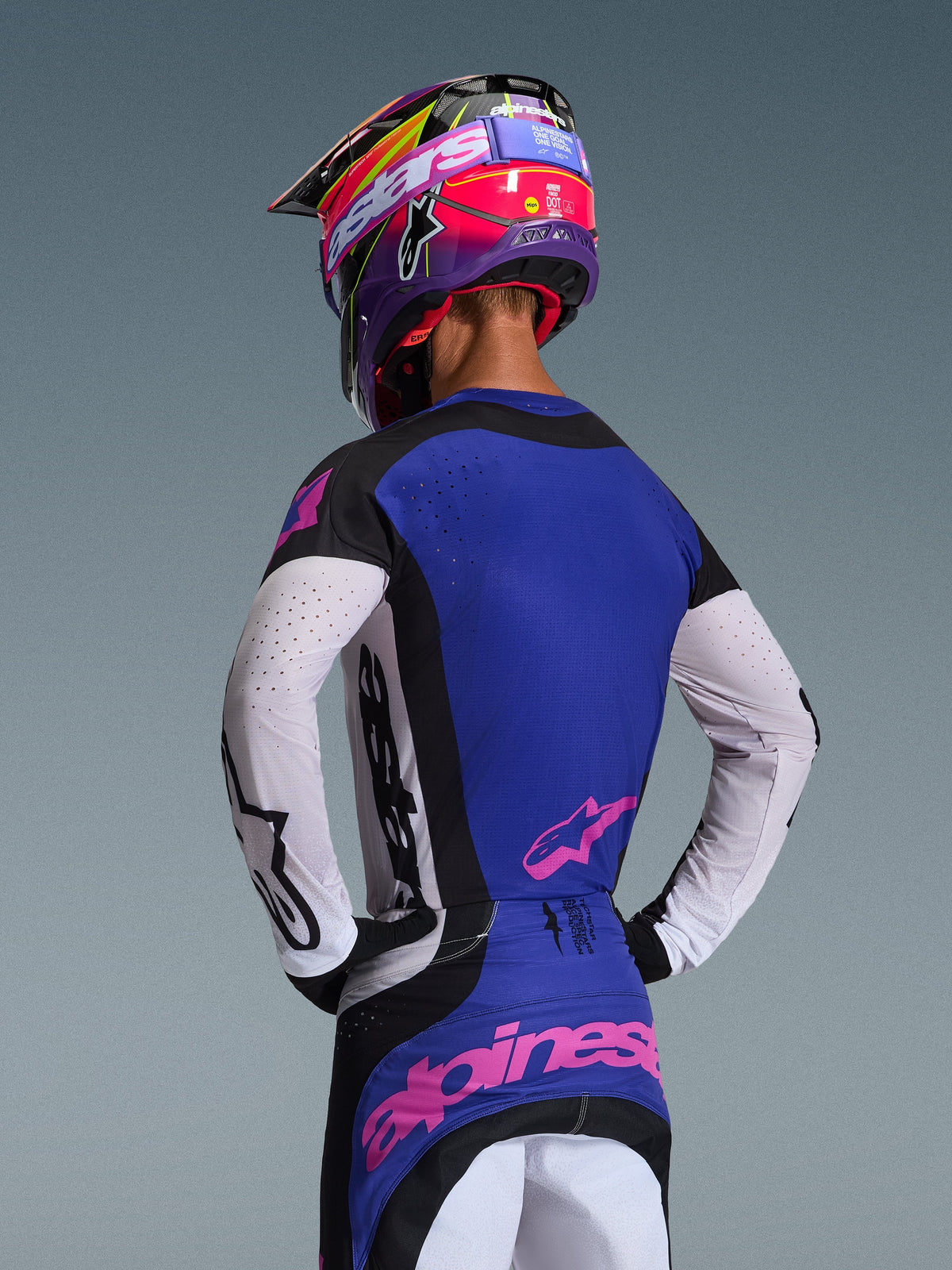 Piloto de motocross visto desde atrás vistiendo el Jersey Techstar Knif 2026, en negro, gris claro y morado, con mangas blancas perforadas y logotipos de Alpinestars en rosa, combinado con un casco morado y rosa a juego, equipamiento profesional de carreras off-road.