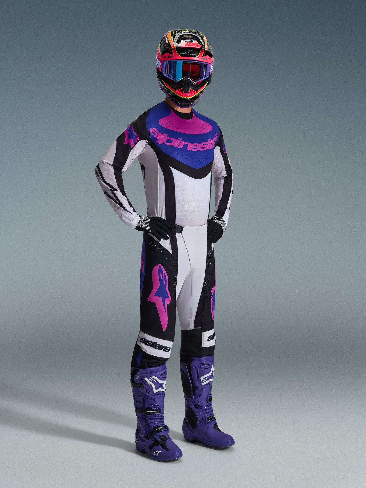 Piloto de motocross con el Jersey Alpinestars Techstar Knif 2026 en Negro Gris Claro Púrpura, pantalones a juego con logotipos rosas grandes, botas púrpuras y casco multicolor con gafas azules, equipamiento completo de carreras para rendimiento off-road