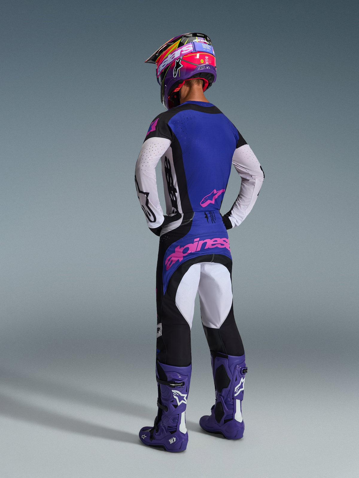 Piloto de motocross visto desde atrás con equipamiento de carreras completo, incluyendo el Jersey Techstar Knif 2026 con espalda azul y mangas blancas perforadas, pantalones negros a juego con logotipos de Alpinestars en rosa, casco multicolor y botas moradas, conjunto coordinado para competición off-road.