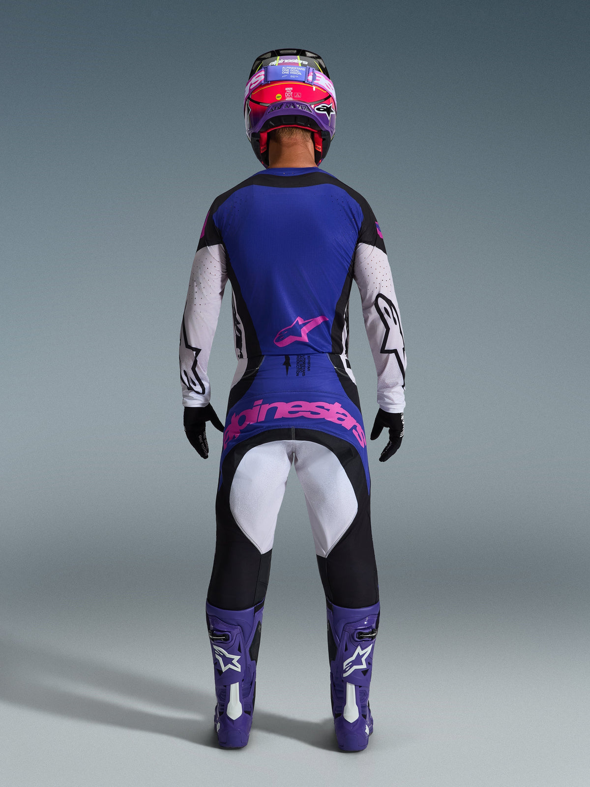 Piloto de motocross visto desde atrás usando el Jersey Techstar Knif 2026, negro, gris claro y morado, pantalones de motocross a juego con marca en rosa, botas y casco morados, equipamiento coordinado de alto rendimiento para off-road