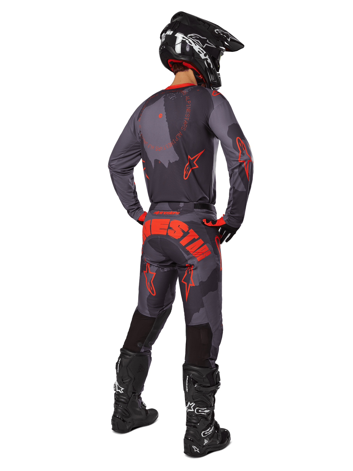Piloto de motocross visto desde atrás con el jersey Alpinestars 2025 Racer Hollow en gris camuflaje y naranja flúor, pantalones de camuflaje a juego con texto de Alpinestars en naranja, casco negro y botas negras, conjunto de equipamiento coordinado para conducción off-road