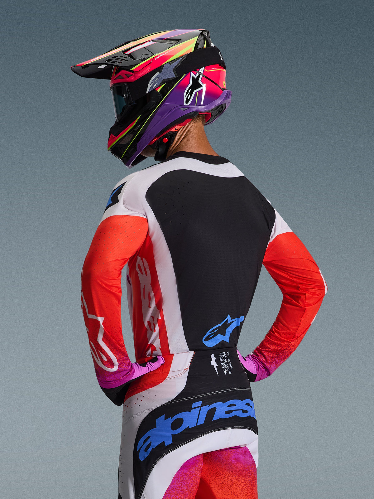 Alpinestars Techstar Knif Jersey 2026, jersey de motocross visto desde atrás en gris claro, naranja flúor y negro, con panel trasero negro con ventilación láser, mangas naranjas y paneles laterales grises, combinado con pantalones a juego y un casco colorido.