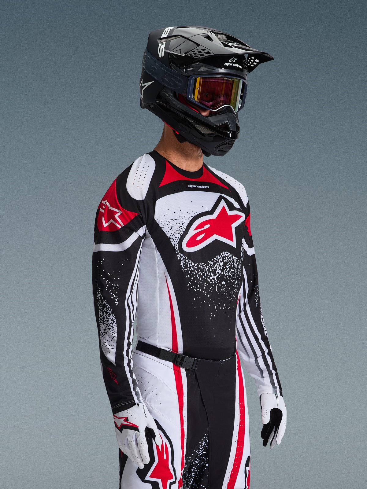 Piloto de motocross con el Jersey Alpinestars Techstar Nomur 2026 en color negro, blanco y rojo, con un gran logotipo Astars rojo y patrones gráficos moteados, combinado con pantalones de carreras a juego, guantes blancos y casco negro con gafas, conjunto coordinado para competición todoterreno