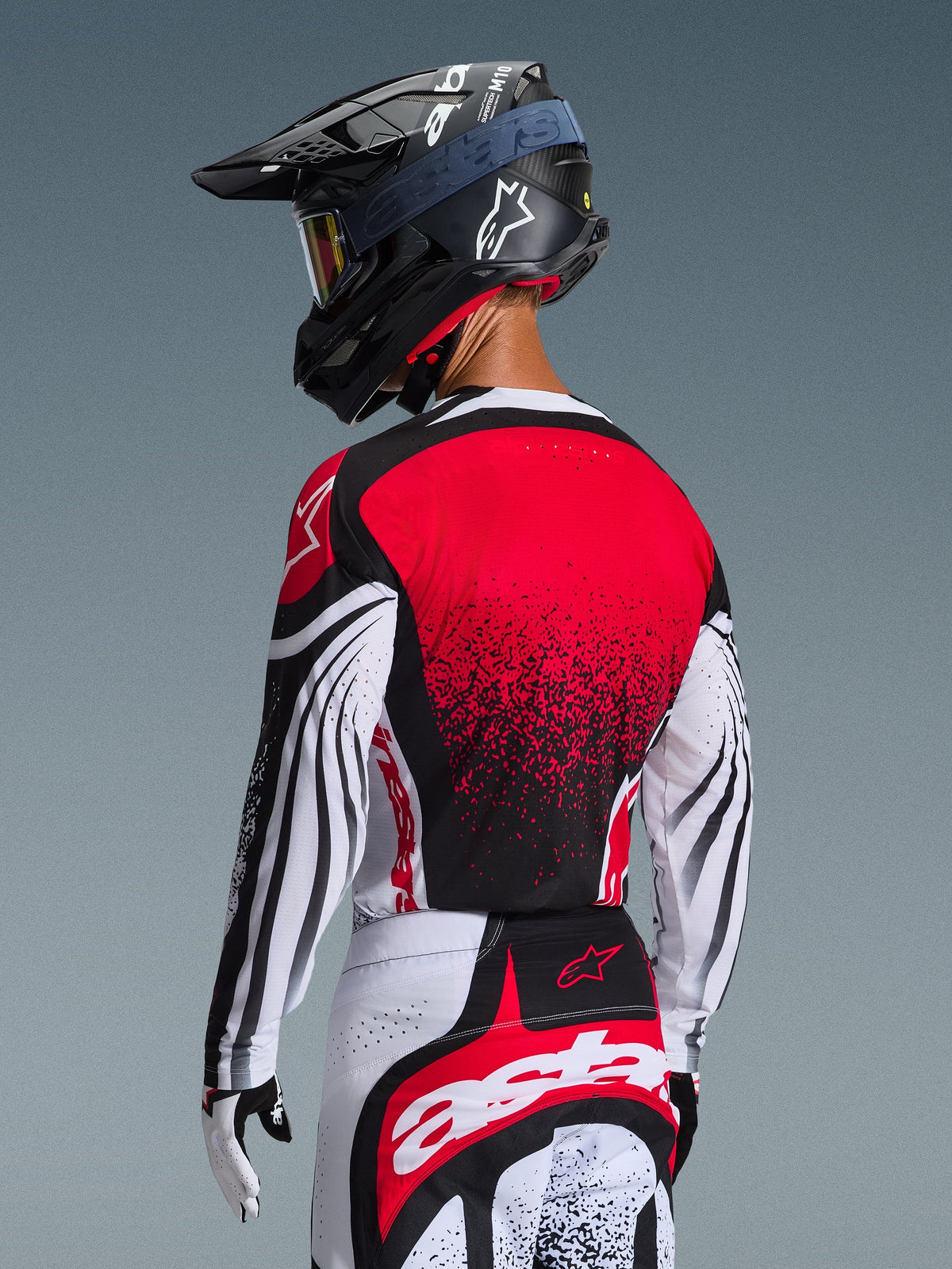 Piloto de motocross desde atrás con el Jersey Techstar Nomur 2026, espalda roja con diseño de degradado negro y mangas blancas con detalles negros, combinado con pantalones rojos y blancos a juego, casco negro y guantes blancos, equipamiento profesional de carreras todoterreno