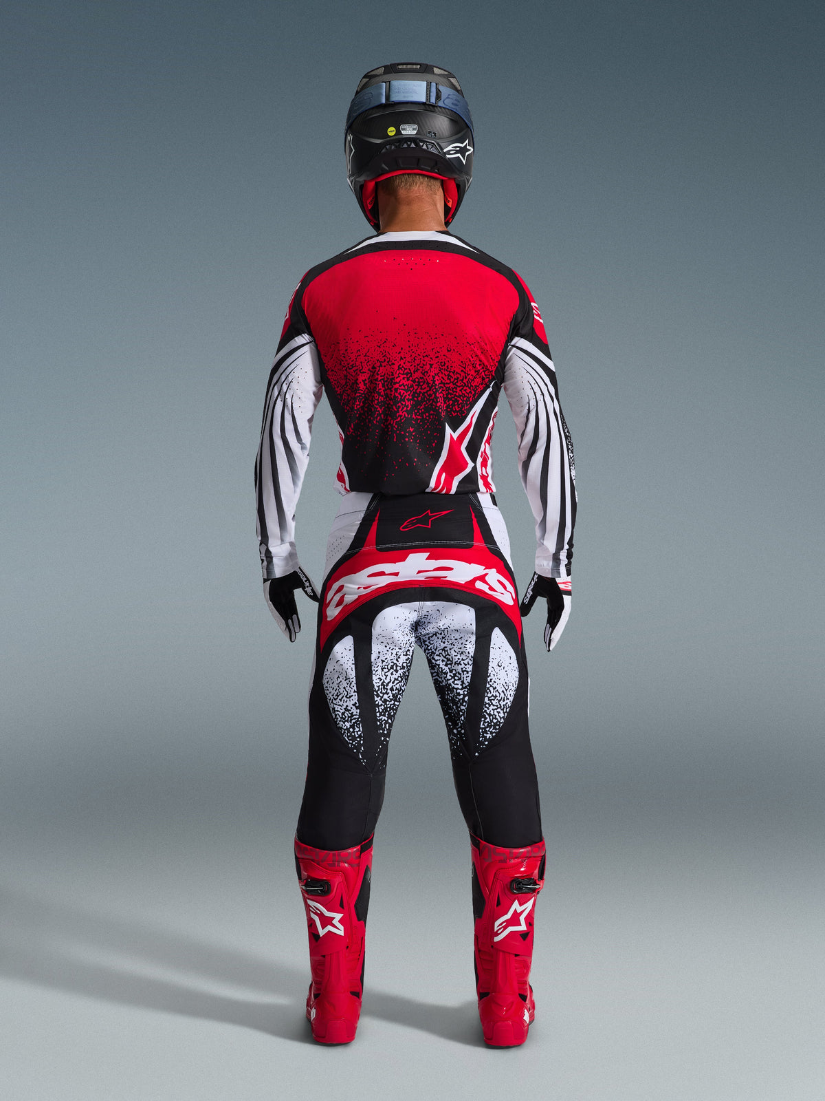 Motocross rider from behind wearing the Alpinestars 2026 Techstar Nomur Gear Set, red and white jersey with black speckle gradient, matching white and red pants with black accents, bright red boots, and black helmet, coordinated off-road racing gear.