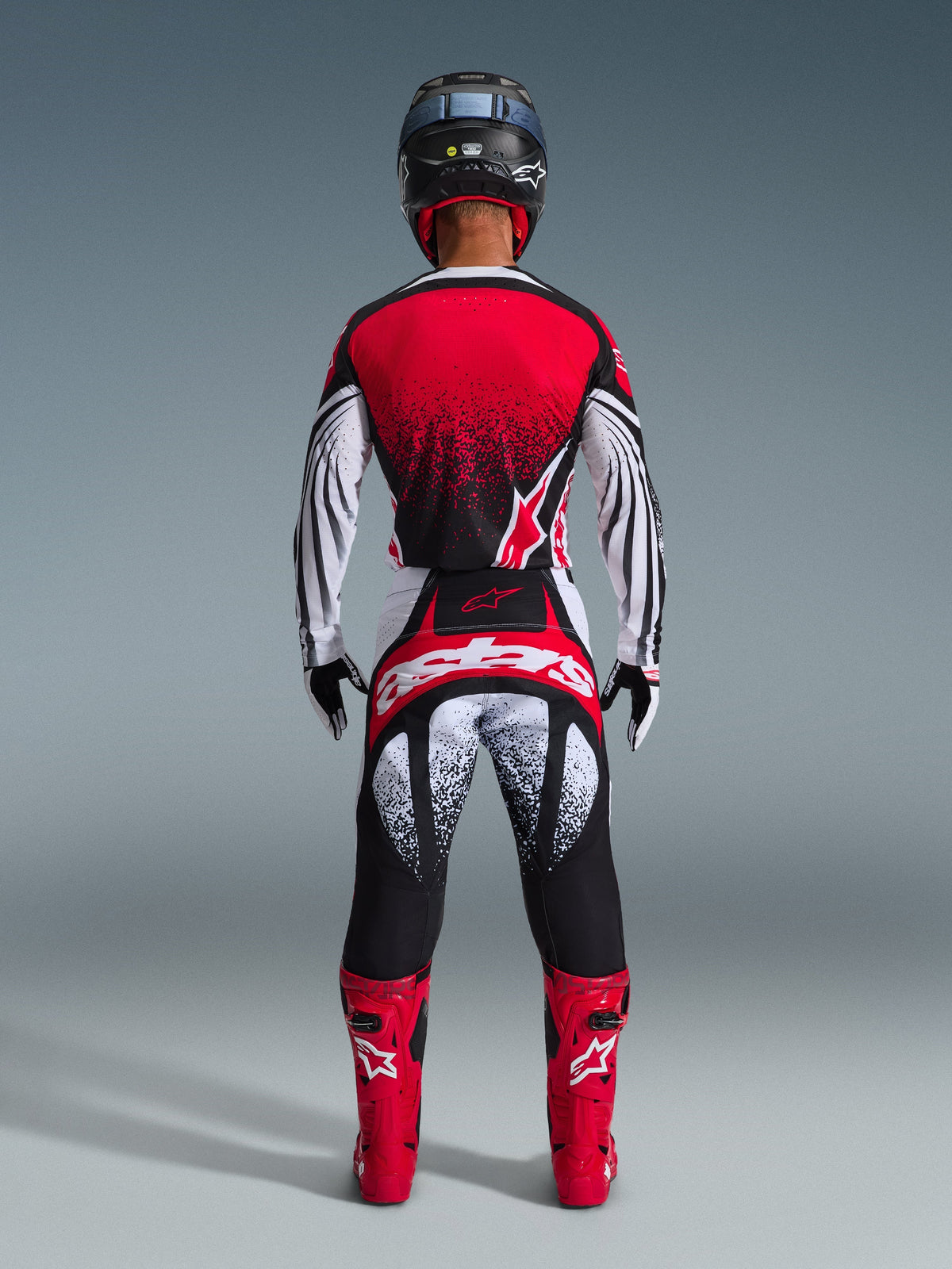 Piloto de motocross desde atrás vistiendo el Jersey Alpinestars Techstar Nomur 2026, rojo, negro y blanco con patrón punteado, pantalones de carreras a juego, botas de motocross rojas y casco negro, conjunto completo de equipamiento para competición off-road