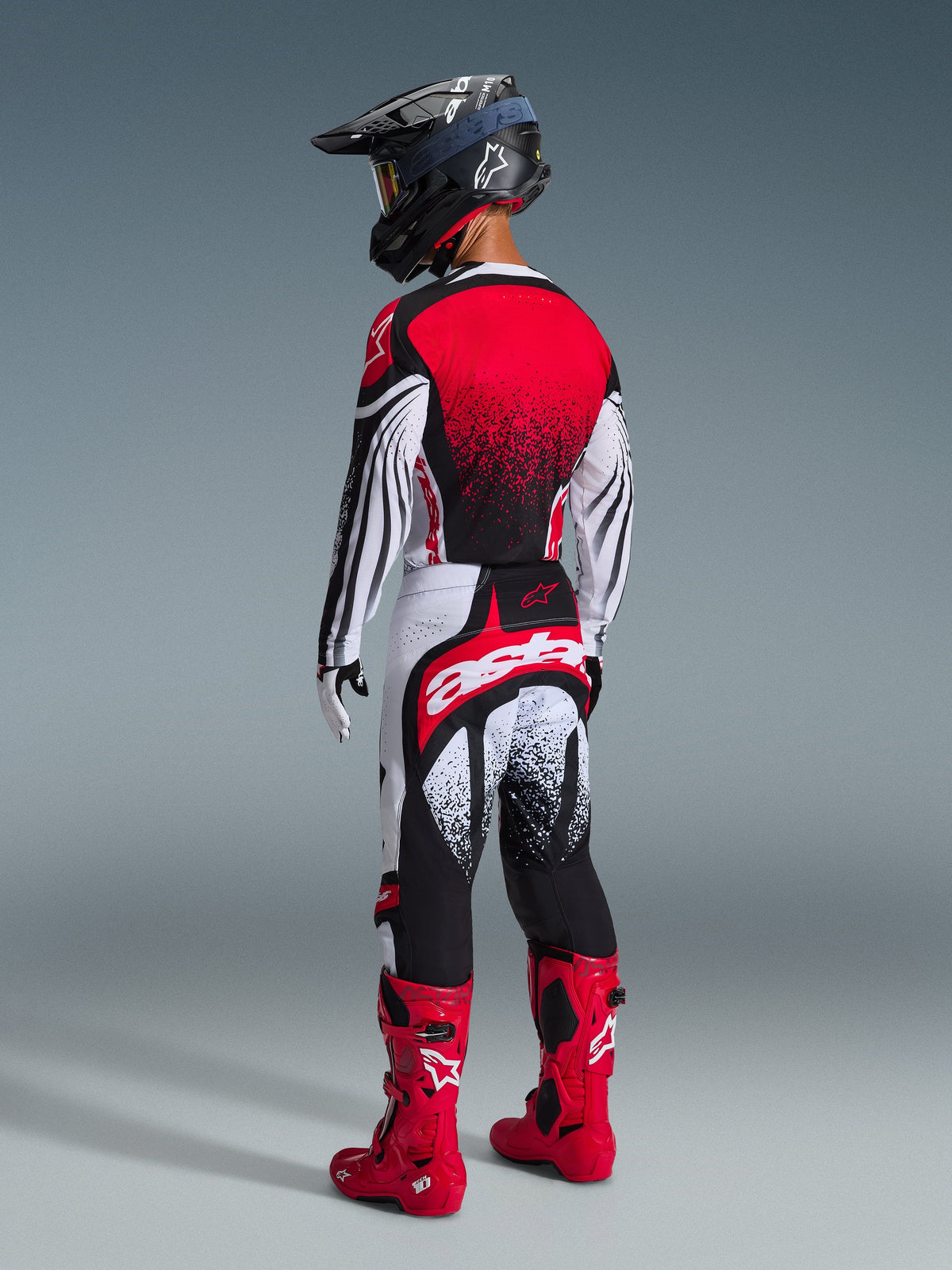 Vista trasera de un piloto de motocross que viste el Jersey Alpinestars Techstar Nomur 2026 en negro, blanco y rojo, con un degradado rojo en la espalda y patrones ondulados en las mangas, combinado con pantalones de motocross a juego, botas rojas y un casco negro para un conjunto de competición off-road coordinado.