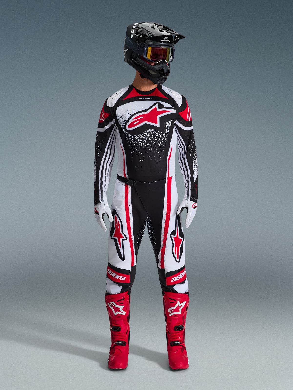 Piloto de motocross con equipamiento completo de Alpinestars, Jersey Techstar Nomur 2026 en negro, blanco y rojo, pantalones a juego con detalles rojos, casco negro y botas rojas brillantes, con un gran logotipo Astars rojo en el pecho y patrón de degradado moteado, conjunto coordinado para competición todoterreno