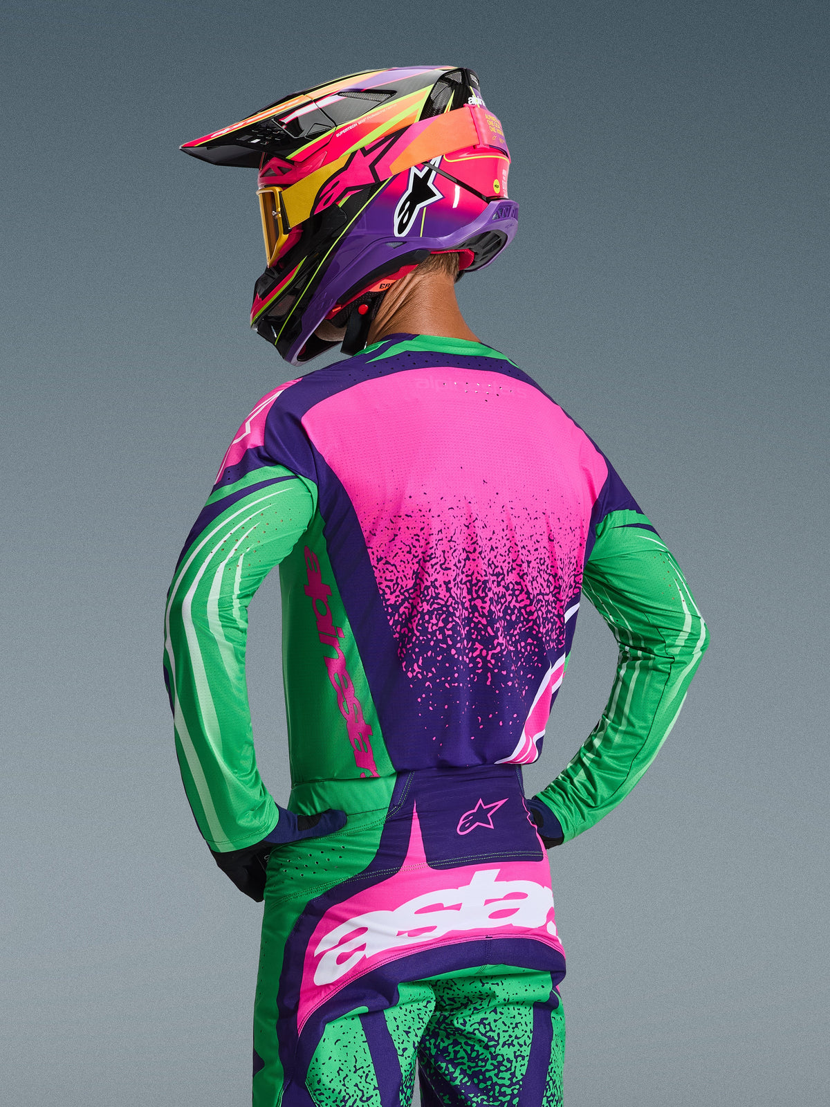 Piloto de motocross visto desde atrás con el conjunto de equipo Alpinestars 2026 Techstar Nomur, jersey y pantalones a juego en púrpura oscuro, verde y rosa flúor, con gráficos llamativos y un ajuste de carrera atlético, combinado con un casco multicolor y gafas amarillas.