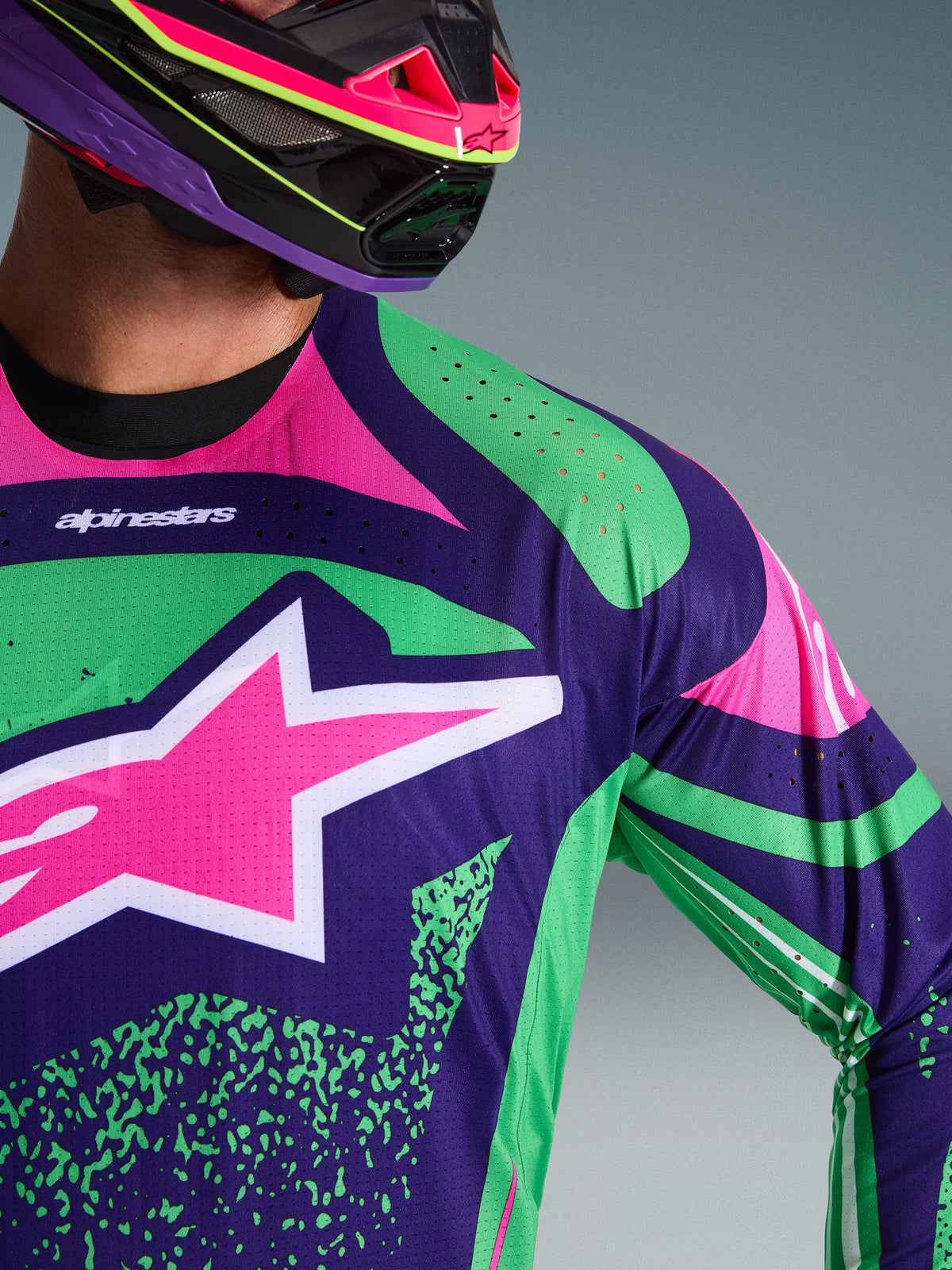 Alpinestars Techstar Nomur Jersey 2026, jersey de motocross en color púrpura intenso con detalles en verde y rosa flúor, con un gran logotipo de estrella rosa en el pecho y perforaciones cortadas con láser en el hombro, usado por un piloto con casco de motocross a juego en púrpura y rosa