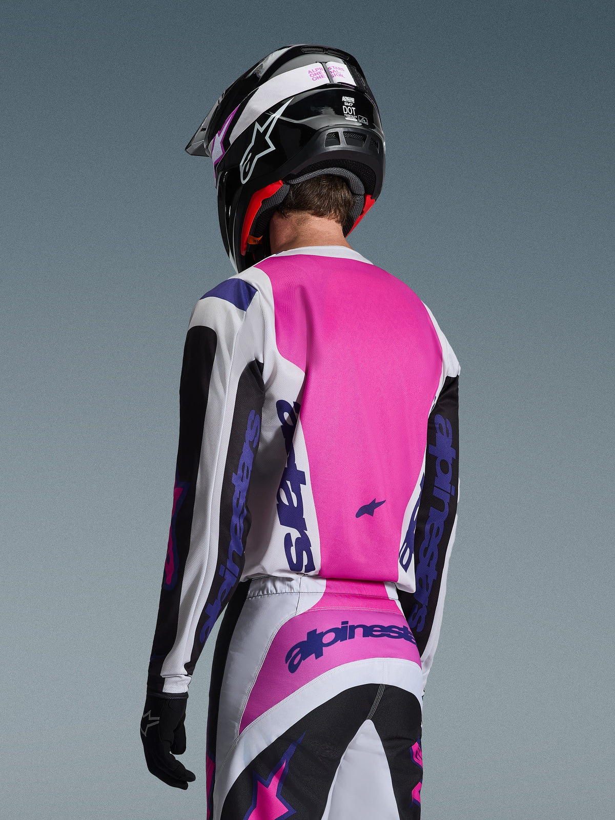 Piloto de motocross visto desde atrás vistiendo el Jersey Alpinestars Fluid Grid 2026 en negro, blanco y púrpura, pantalones a juego con logotipos llamativos, casco negro con detalles en rosa y guantes negros; equipamiento completo coordinado para el rendimiento off-road.