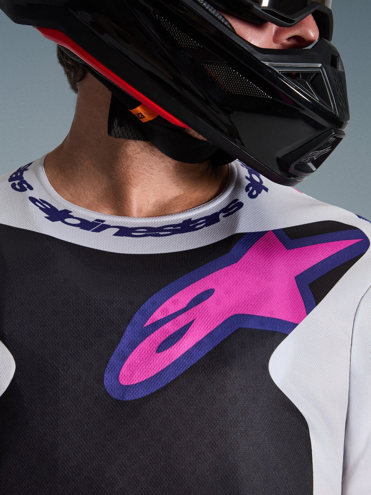Close-up of Alpinestars 2026 Fluid Grid Jersey, Motocross Jersey, Black Light Gray and Purple, featuring a vibrant pink-purple star logo on the chest and branded sport collar, worn with a black motocross helmet