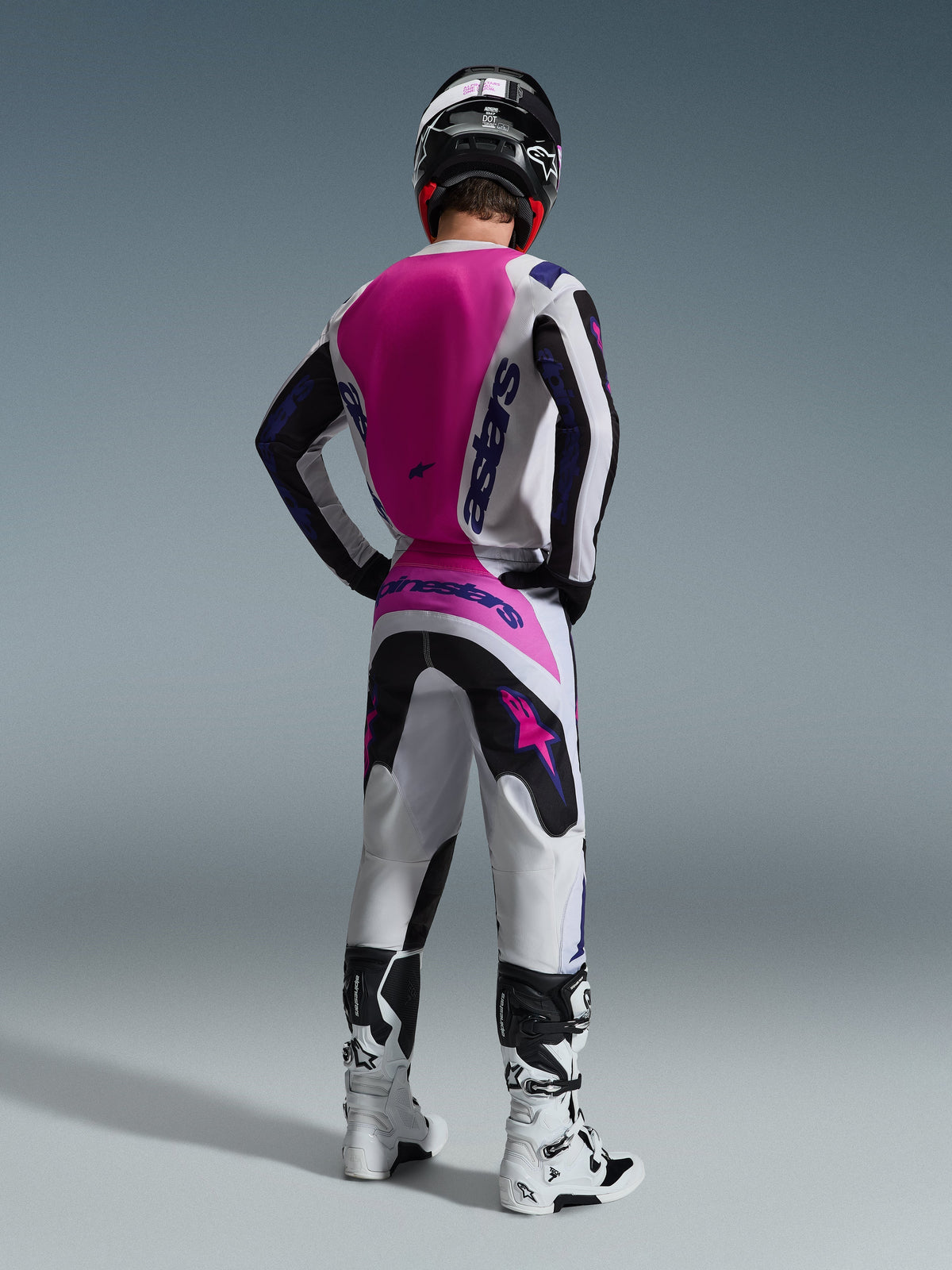 Alpinestars 2026 Fluid Grid Gear Set, Motocross rider seen from the back, white jersey with a pink back panel and purple branding, matching white and black pants with pink accents, white and black boots, and a black helmet, coordinated off-road racing outfit.
