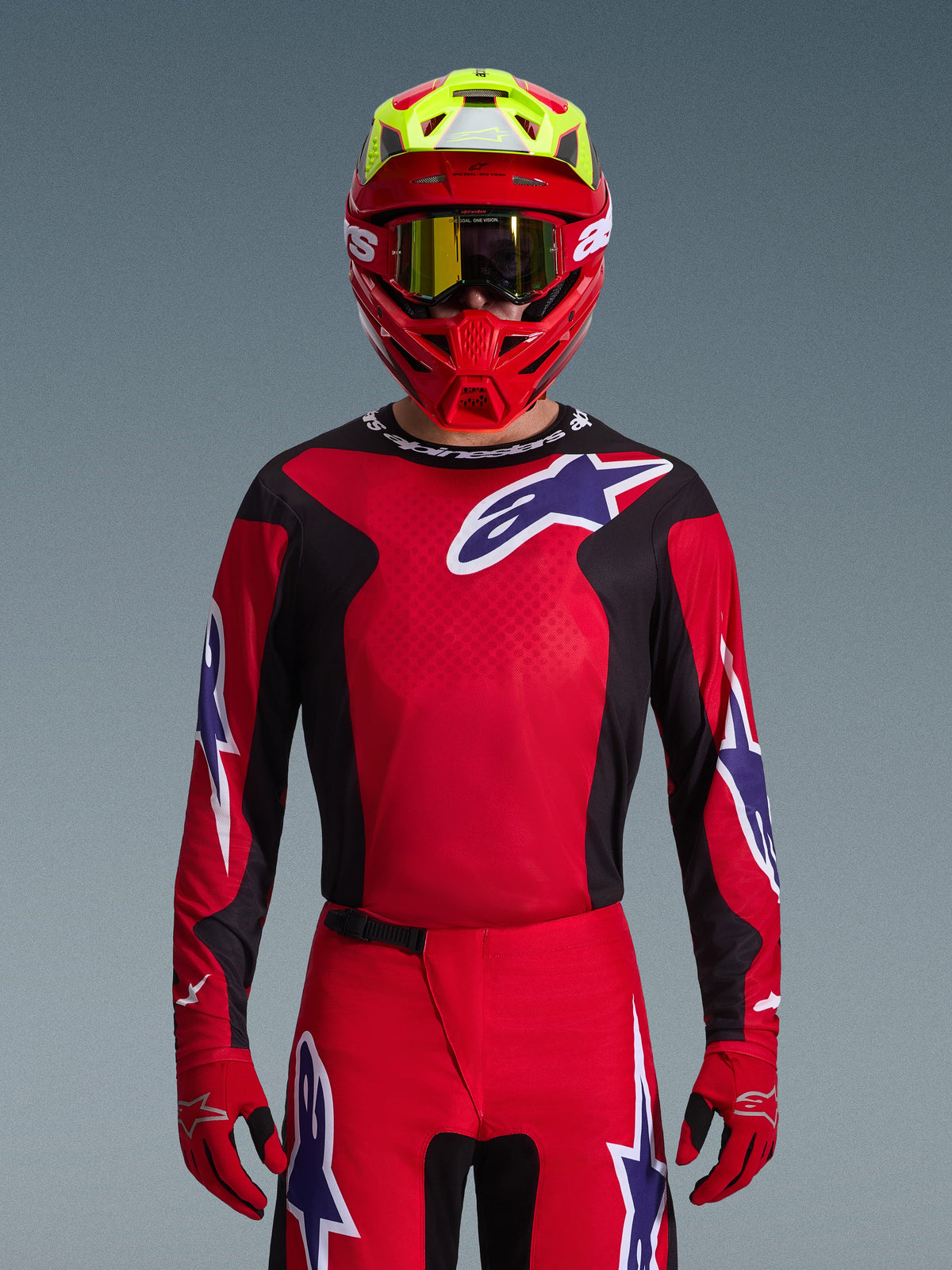 Piloto de motocross con el conjunto Alpinestars Fluid Grid 2026, jersey rojo con paneles laterales negros y logo de estrella blanco, pantalones y guantes rojos a juego, casco rojo y amarillo con gafas de lente dorada, equipamiento de carreras de alto rendimiento para off-road
