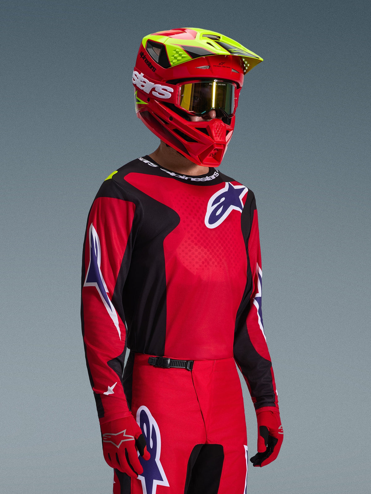 Piloto de motocross con el jersey Alpinestars Fluid Grid 2026 en rojo, negro y púrpura, combinado con pantalones de motocross a juego, casco rojo con detalles en amarillo neón, gafas doradas y guantes rojos, conjunto completo de carreras off-road.