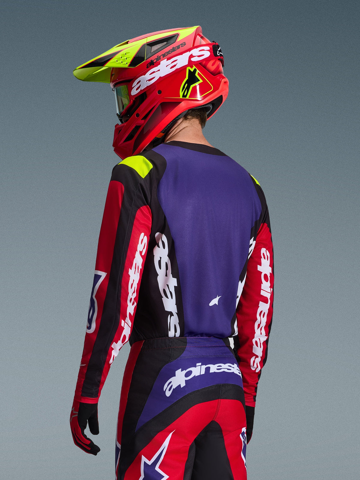 Piloto de motocross con el Jersey Alpinestars Fluid Grid 2026, rojo, negro y púrpura, vista trasera que muestra el panel posterior púrpura y las mangas rojas con logotipos blancos, pantalones a juego y un casco rojo y amarillo neón, conjunto coordinado para carreras de motocross