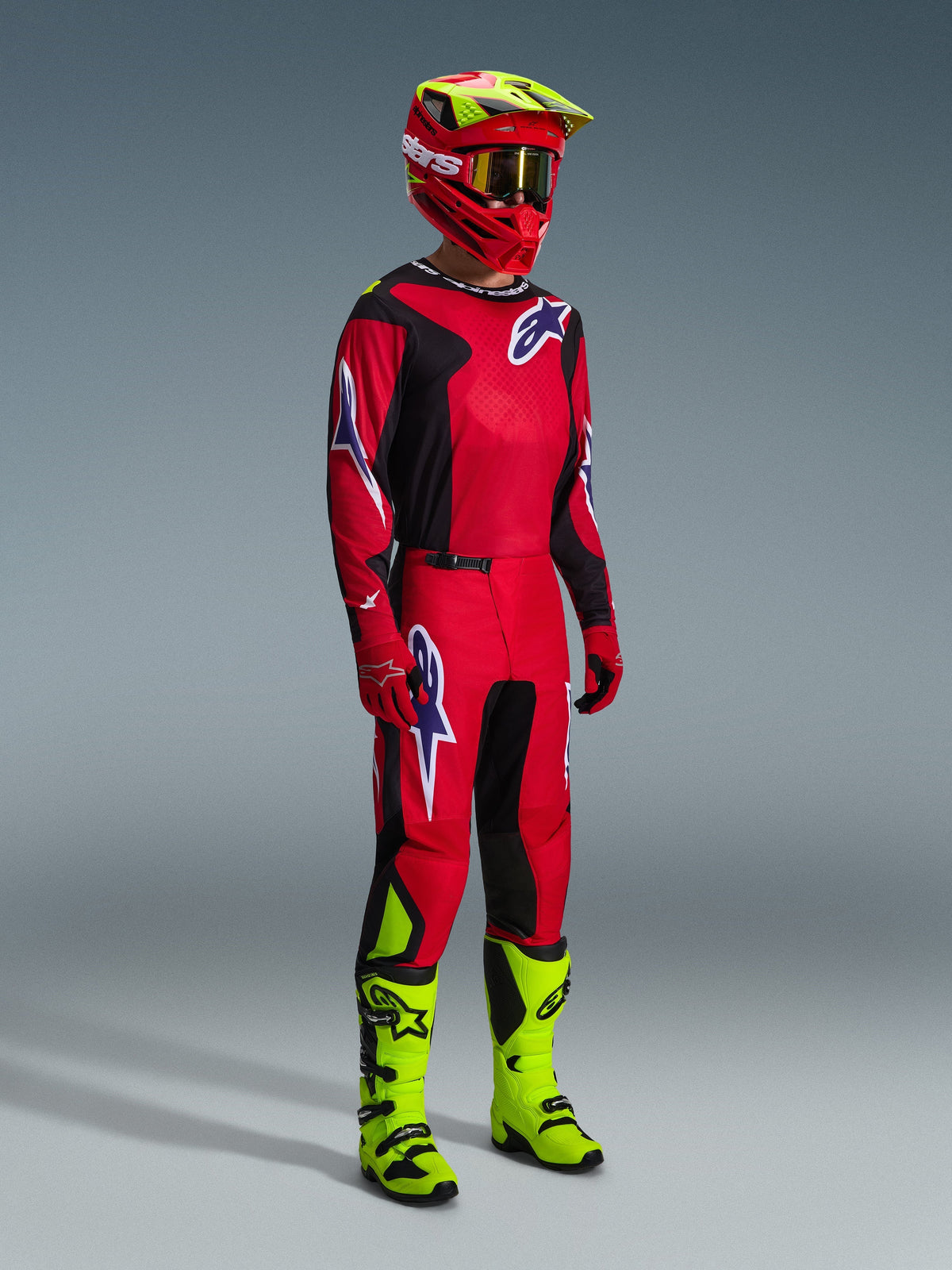 Piloto de motocross luciendo el conjunto Alpinestars 2026 Fluid Grid en rojo, negro y morado; incluye jersey y pantalones con logotipos morados, casco rojo y amarillo neón, y botas amarillo neón para un kit completo de carreras off-road.