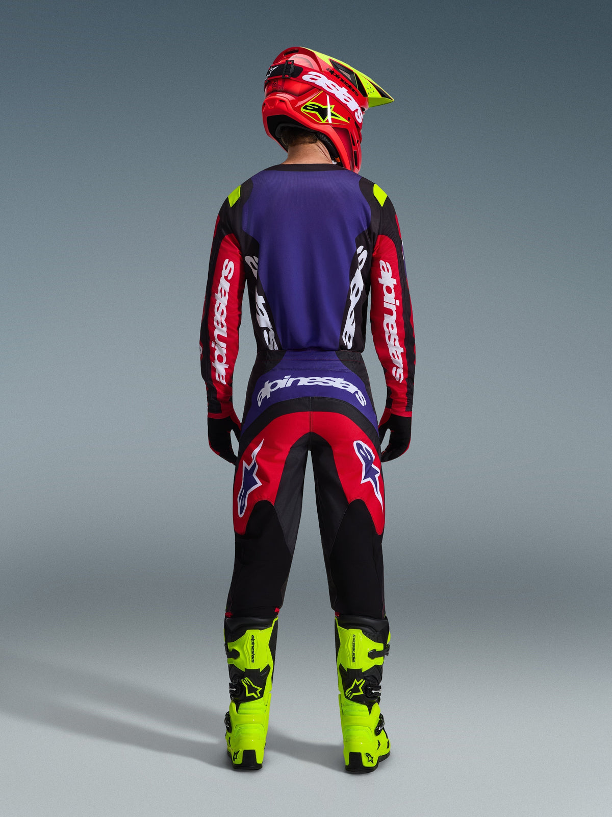 Piloto de motocross visto desde atrás con el conjunto Alpinestars 2026 Fluid Grid en rojo, negro y púrpura, con un panel trasero púrpura y mangas rojas en el jersey, pantalones rojos y negros a juego, botas de motocross amarillo neón brillante y un casco rojo y amarillo neón.