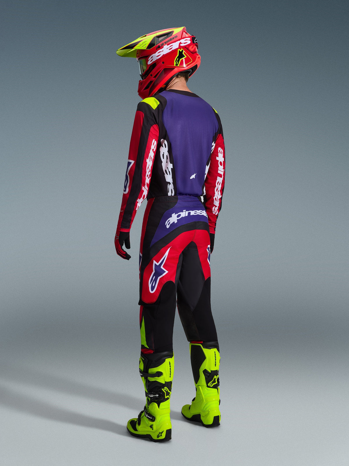 Piloto de motocross con el conjunto Alpinestars 2026 Fluid Grid visto desde atrás, jersey rojo, negro y morado con marca en blanco, pantalones rojos y negros con detalles morados, combinado con casco rojo y amarillo neón y botas amarillo neón brillante, equipamiento completo de carreras off-road
