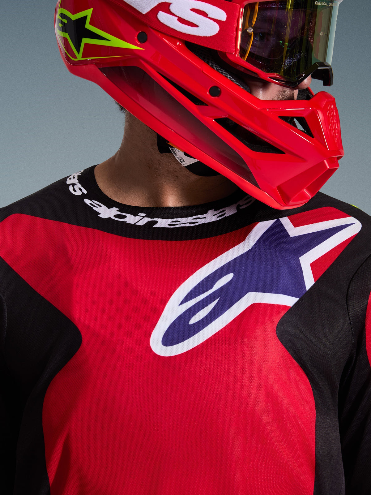 Primer plano de un piloto de motocross con el Jersey Fluid Grid 2026 en rojo, negro y púrpura, con un gran logotipo de Alpinestars en el pecho y cuello deportivo con marca, combinado con casco rojo y gafas, destacando el tejido técnico de polifibra y el ajuste atlético.
