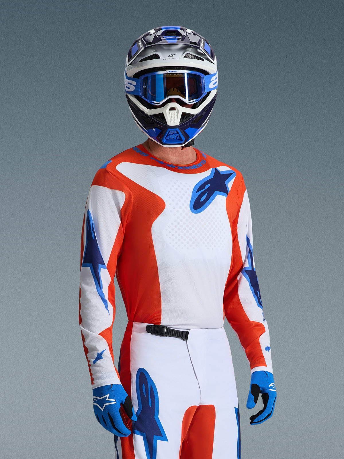 Piloto de motocross con el conjunto Alpinestars 2026 Fluid Grid, jersey naranja y blanco con logotipos de estrellas azules, pantalones blancos y naranjas con acentos azules, guantes azules, casco azul y blanco con gafas azules, equipamiento deportivo coordinado para alto rendimiento en pista