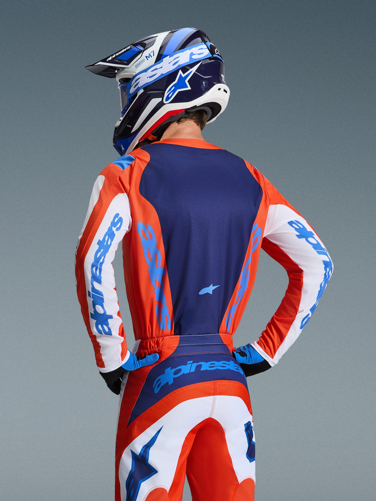 Alpinestars 2026 Fluid Grid Jersey, vista posterior de un piloto de motocross con conjunto coordinado en naranja y azul UCLA, panel trasero azul marino, detalles blancos en las mangas con marca en azul, casco a juego, ropa técnica para todoterreno